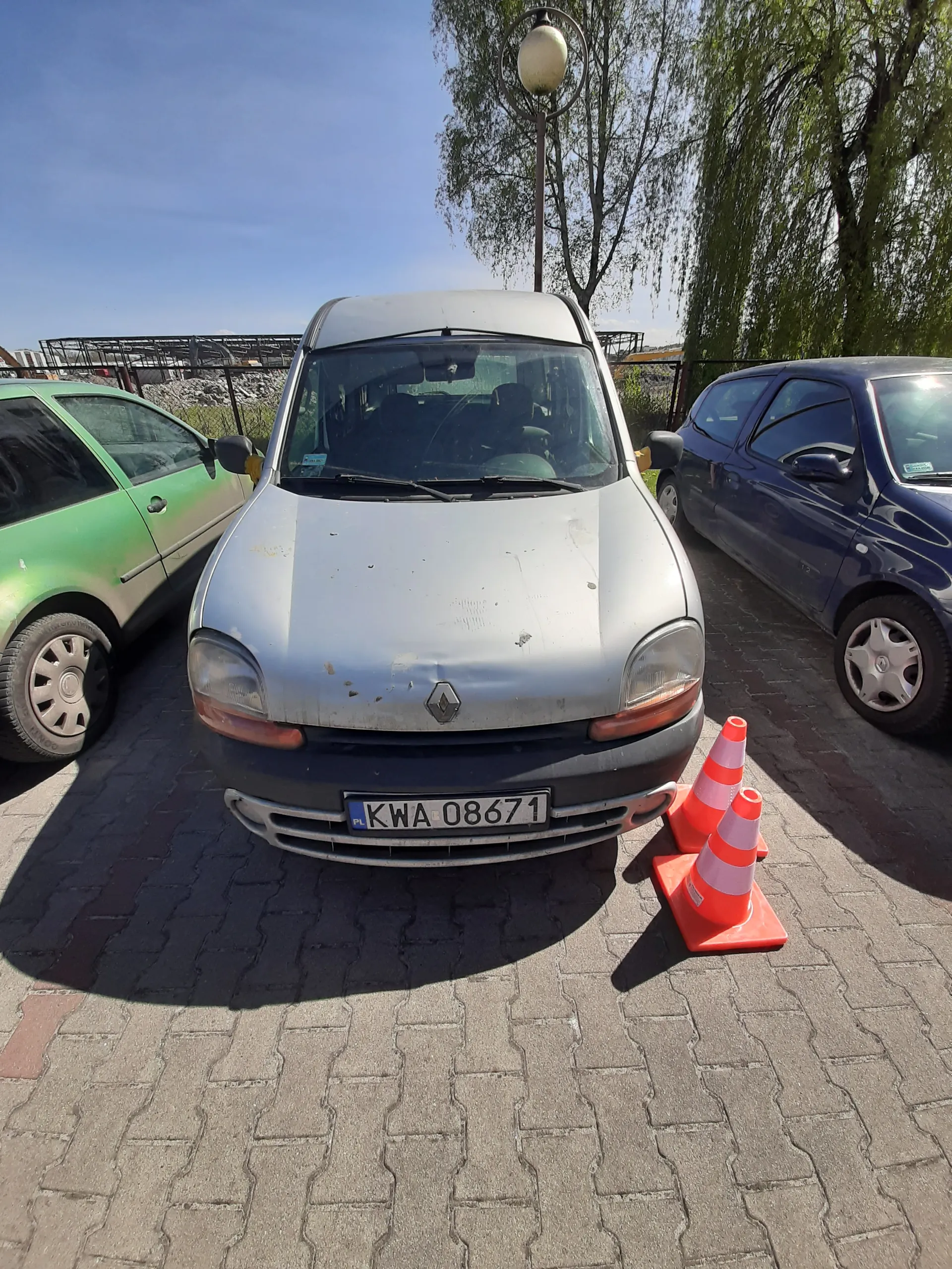 Trzy samochody zaparkowane na brukowanej powierzchni: srebrny Renault z tablicą rejestracyjną KWA 08671, niebieski samochód oraz zielony samochód. Obok srebrnego Renault znajdują się dwa pomarańczowe stożki drogowe. W tle widoczne są drzewa, latarnia uliczna i błękitne niebo.