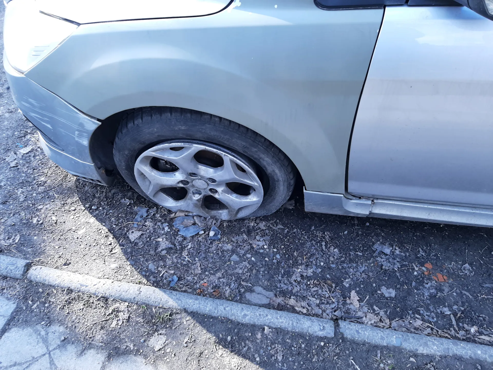 Fragment srebrnego samochodu widziany z boku, z naciskiem na przednie lewe koło z aluminiową felgą. Samochód stoi na nierównym podłożu z drobnymi kamieniami i liśćmi. W dolnej części obrazu widoczny jest betonowy krawężnik. Brak czytelnego tekstu.