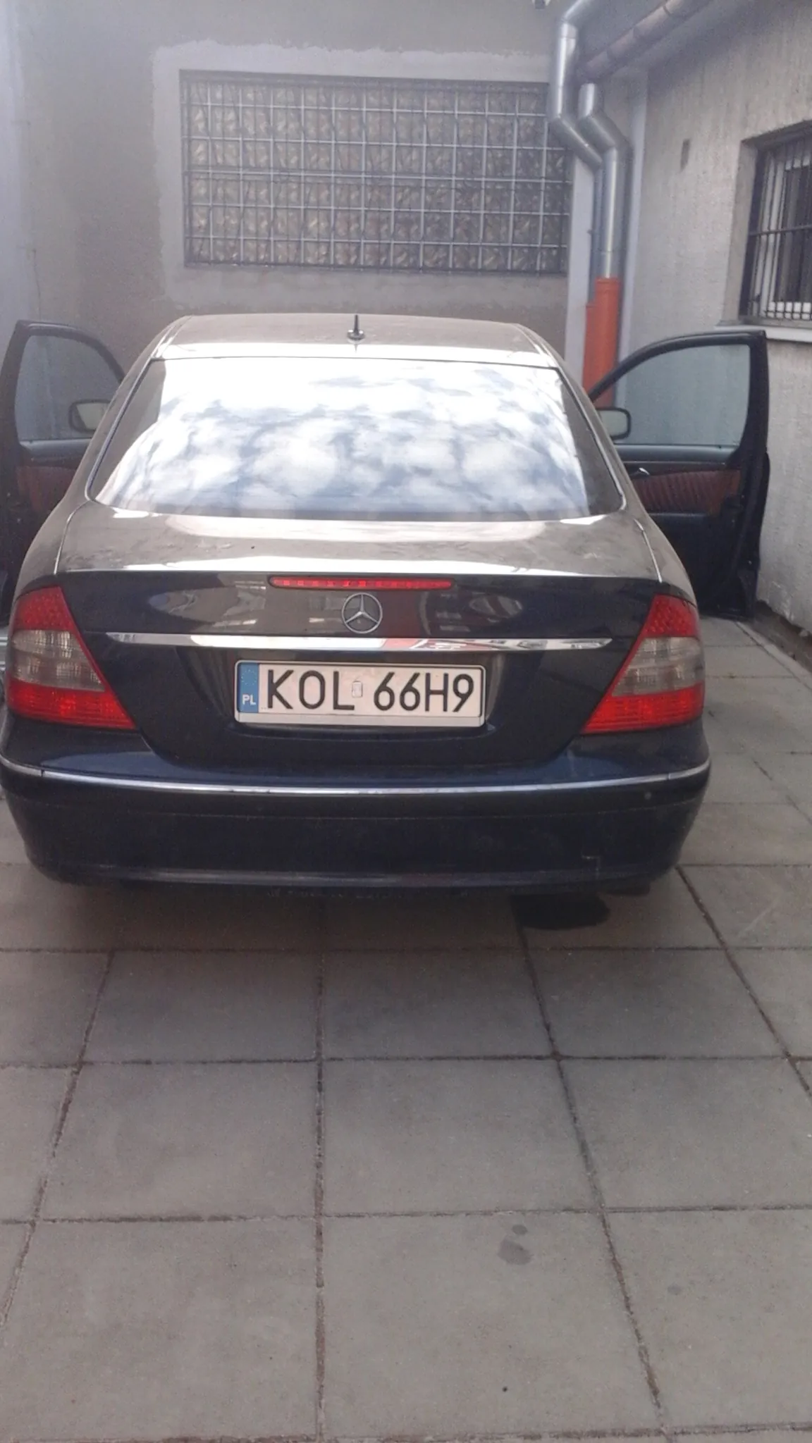 Czarny samochód marki Mercedes-Benz widziany od tyłu z otwartymi drzwiami po obu stronach. Tablica rejestracyjna KOL 66H9. Samochód zaparkowany na chodniku z betonowych płyt obok budynku z oknem zabezpieczonym metalową kratą i rurami. W tylnej szybie odbija się niebo.