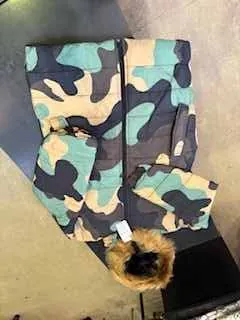 Kurtka w kamuflażu z czarnymi, beżowymi i turkusowymi wzorami, z kapturem wykończonym brązowym futrem. Na kapturze widoczny biały etykietka. Kurtka leży rozłożona na powierzchni, na tle ciemnego i jasnego obszaru, prawdopodobnie w pomieszczeniu.