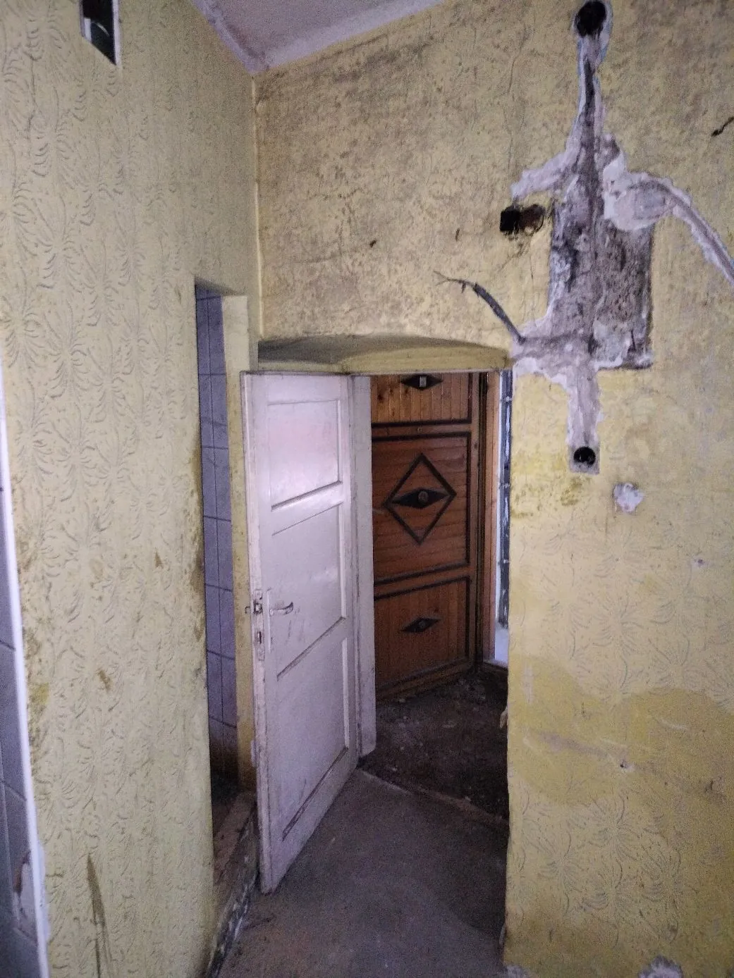 Wnętrze zniszczonego pomieszczenia z żółtymi, chropowatymi ścianami. W centrum znajduje się otwarte, białe drzwi prowadzące do kolejnego pokoju, w którym widoczne są brązowe, drewniane drzwi z rombowym zdobieniem. Na prawej ścianie znajduje się duży, nieregularny ubytek w tynku, odsłaniający szary beton i wystające elementy instalacji.