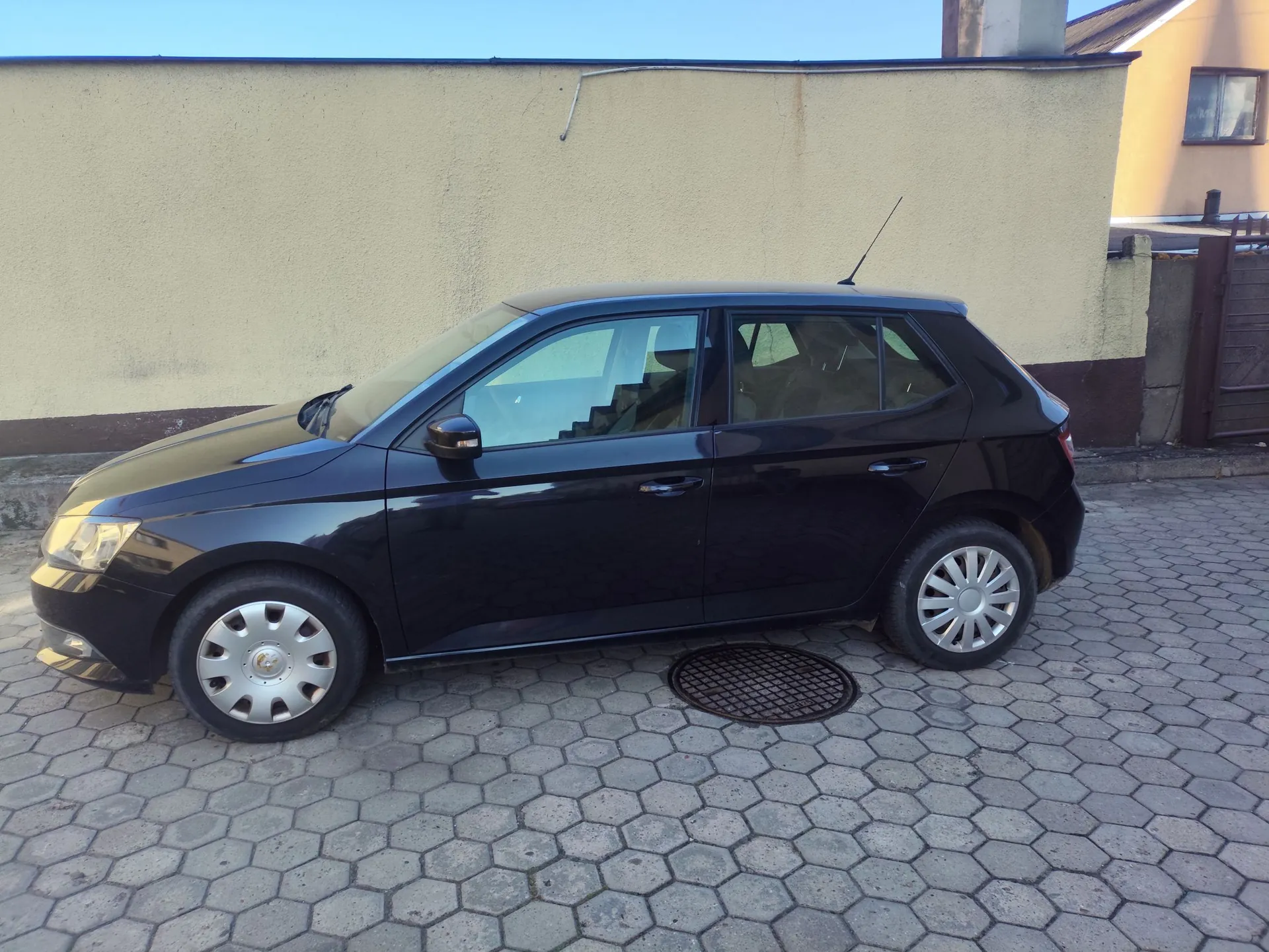Czarny samochód osobowy typu hatchback zaparkowany bokiem na podjeździe z szarej kostki brukowej o sześciokątnym kształcie. W tle znajduje się jasna, gładka ściana budynku oraz fragment żółtej elewacji domu z oknem w prawym górnym rogu. Na ziemi, pod samochodem, widoczna jest okrągła pokrywa studzienki kanalizacyjnej.
