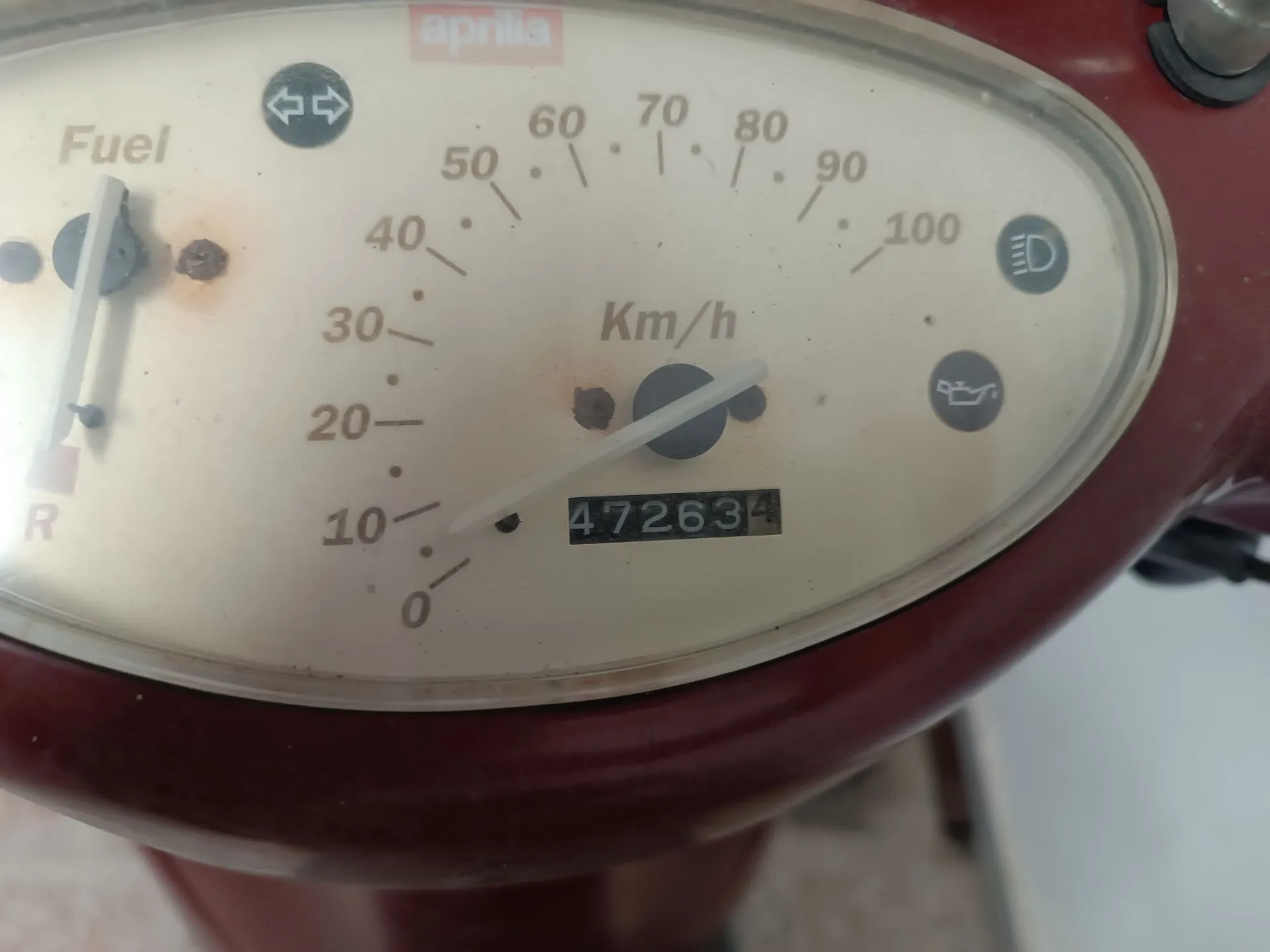 Zbliżenie na licznik prędkości skutera marki Aprilia. Prędkościomierz ma skalę od 0 do 100 km/h, z napisem 'Km/h' w centrum. Wskazówka prędkości znajduje się w okolicach 10 km/h. Poniżej znajduje się licznik przebiegu z wartością 47263. Po lewej stronie widoczny jest wskaźnik poziomu paliwa z napisem 'Fuel' oraz litera 'R'. Po prawej stronie znajdują się dwie ikony kontrolne. Obudowa licznika jest w kolorze czerwonym.