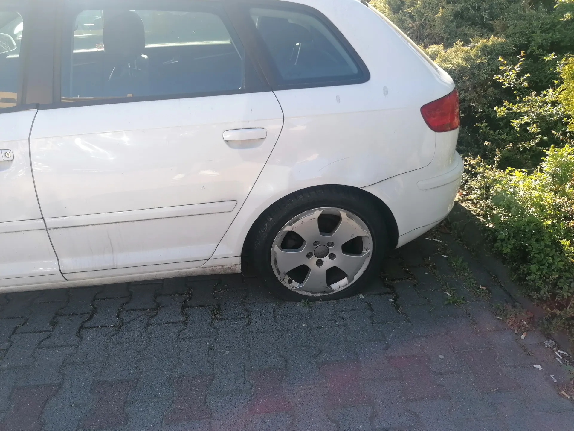 Biały samochód typu hatchback zaparkowany na brukowanej powierzchni. Widoczna tylna część pojazdu z czerwonym światłem tylnym, srebrną felgą i czarną oponą. Po prawej stronie zielone krzewy. Brak czytelnego tekstu.
