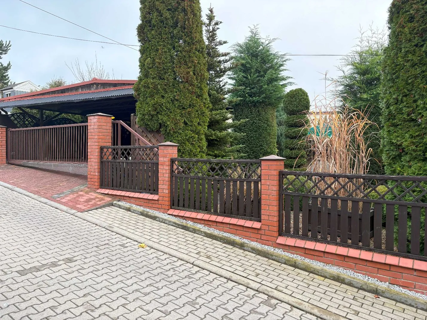Zdjęcie przedstawia fragment posesji z ceglanym niskim murem i ciemnobrązowym drewnianym ogrodzeniem. Za ogrodzeniem rosną wysokie, gęste drzewa iglaste oraz ozdobne trawy. Po lewej stronie widoczna jest wiata z ciemnym dachem i bramą wjazdową. Przed murem znajduje się chodnik wyłożony szarą kostką brukową, a bezpośrednio przy ogrodzeniu czerwona kostka.