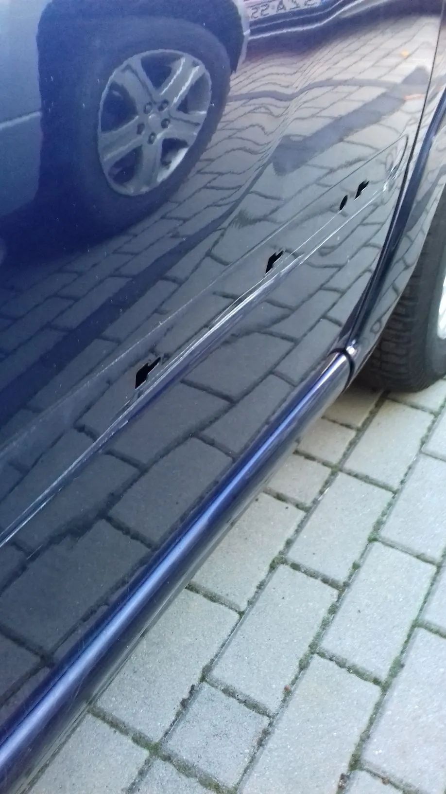 Zbliżenie na ciemnoniebieski lakier samochodu z widocznymi uszkodzeniami w postaci kilku głębokich zarysowań i wgnieceń wzdłuż boku nadwozia. W lakierze odbija się kostka brukowa oraz koło innego pojazdu. Samochód stoi na szarym chodniku z kostki brukowej.
