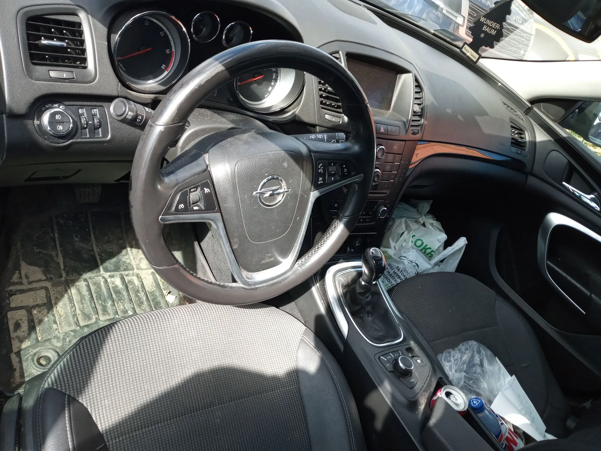 Wnętrze samochodu marki Opel widziane z perspektywy kierowcy. Widoczna jest czarna kierownica z logo producenta w centrum, deska rozdzielcza z analogowymi wskaźnikami prędkości i obrotów oraz konsola środkowa z lewarkiem zmiany biegów. Na siedzeniu pasażera i w przestrzeni obok leżą białe plastikowe torby. Na podłodze znajduje się gumowa mata. W prawym dolnym rogu widoczna jest puszka i butelka w schowku.