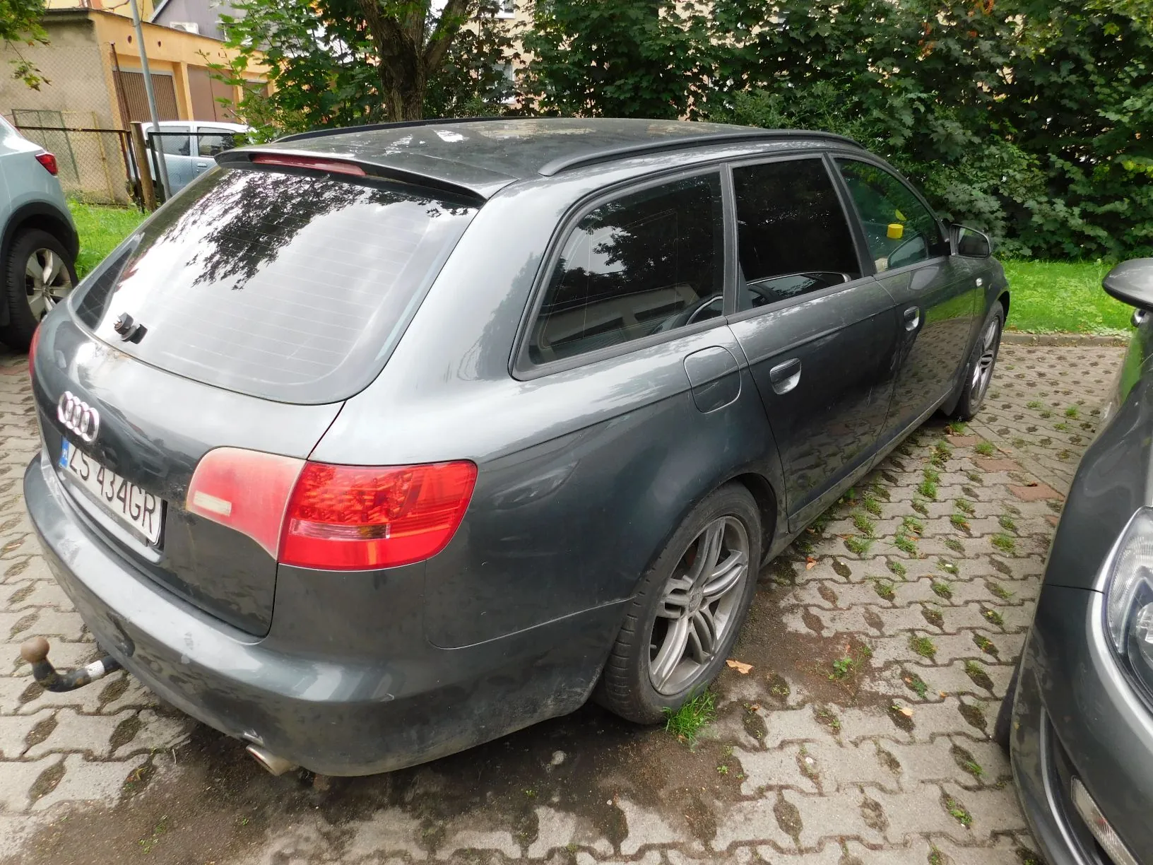 Szary samochód marki Audi w nadwoziu kombi, zaparkowany na kostce brukowej. Widoczny jest tył i prawy bok pojazdu. Samochód ma polską tablicę rejestracyjną o numerze ZS 434GR oraz zamontowany hak holowniczy. W tle widać zielone krzewy, drzewa oraz fragmenty innych samochodów.
