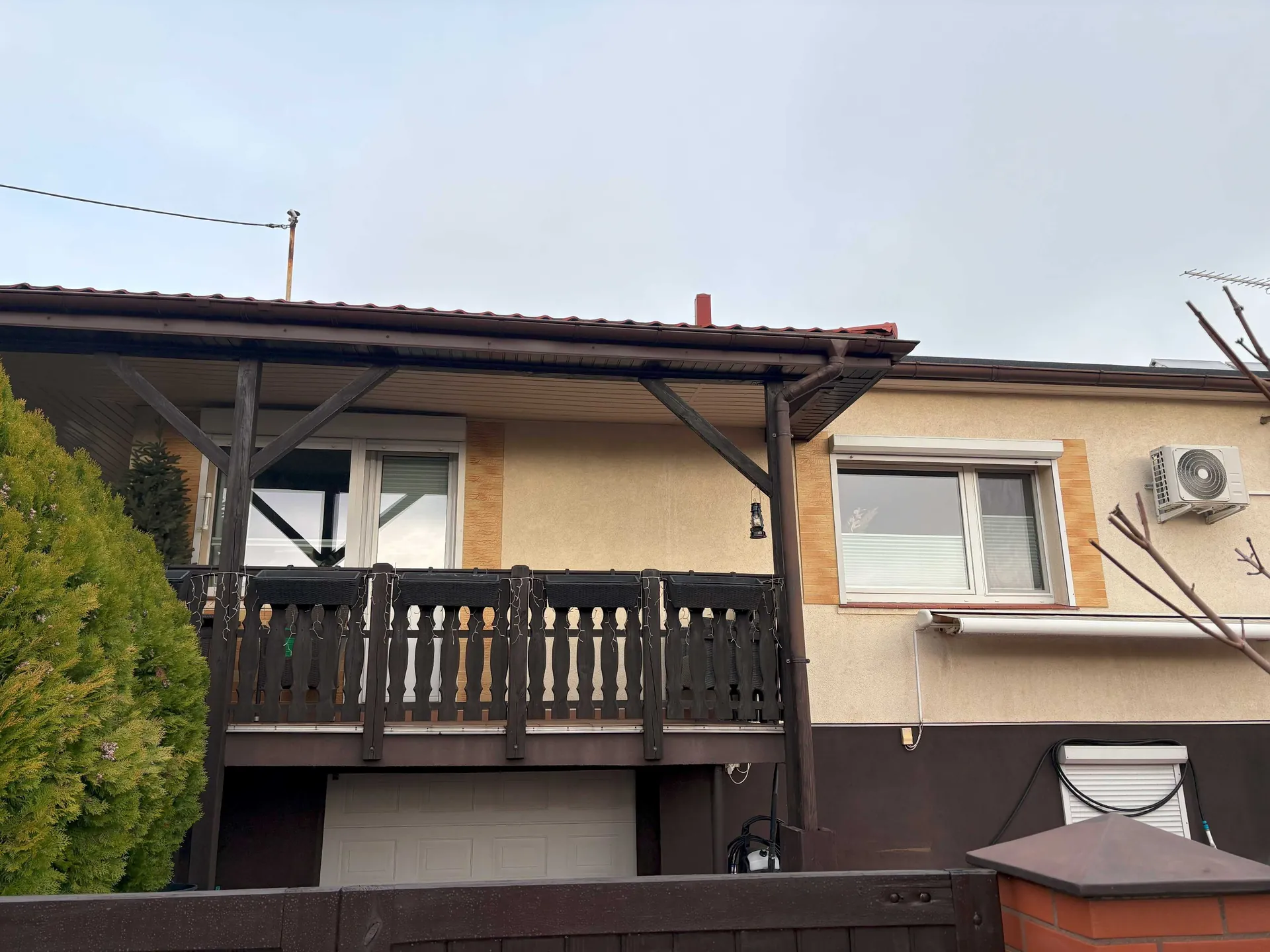 Zdjęcie fragmentu domu jednorodzinnego z balkonom na piętrze. Balkon ma ciemnobrązowe drewniane balustrady i jest przykryty zadaszeniem wspartym na drewnianych słupach. Po prawej stronie znajduje się białe okno z roletą oraz zamontowana na ścianie jednostka zewnętrzna klimatyzatora. Poniżej balkonu widoczna jest biała brama garażowa. Po lewej stronie zdjęcia znajduje się gęsty, zielony krzew iglasty. Ściana domu jest dwukolorowa: jasna w górnej części i ciemnobrązowa w dolnej.