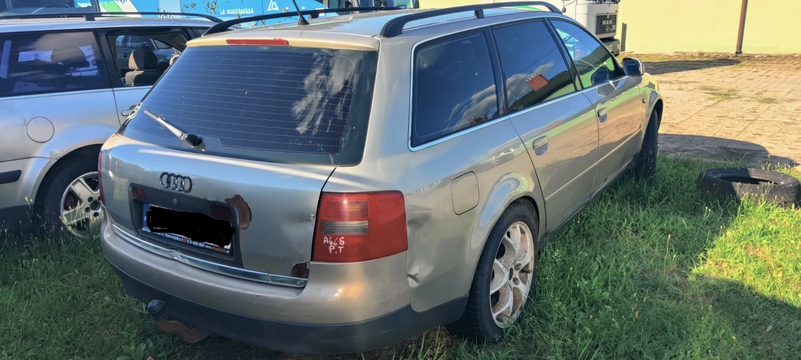 Srebrny samochód marki Audi w nadwoziu kombi, zaparkowany na trawie. Pojazd jest uszkodzony: widoczne jest wgniecenie i odpryski lakieru na tylnym zderzaku oraz klapie bagażnika, a także wgniecenie na tylnym prawym błotniku. Na tylnej lampie widnieje czerwony napis 'A665 P.T.'. W tle widoczny jest fragment innego srebrnego samochodu oraz opona leżąca na trawie.