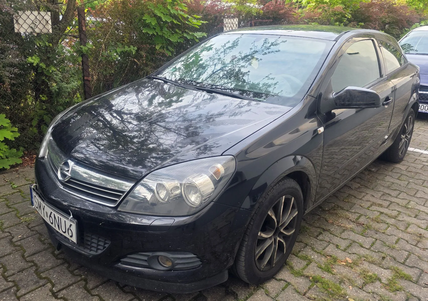 Czarny samochód marki Opel zaparkowany na kostce brukowej. Widoczny jest przód i prawy bok pojazdu. Samochód ma tablicę rejestracyjną o numerze SMY 6NU6. W tle znajdują się zielone krzewy i drzewa.
