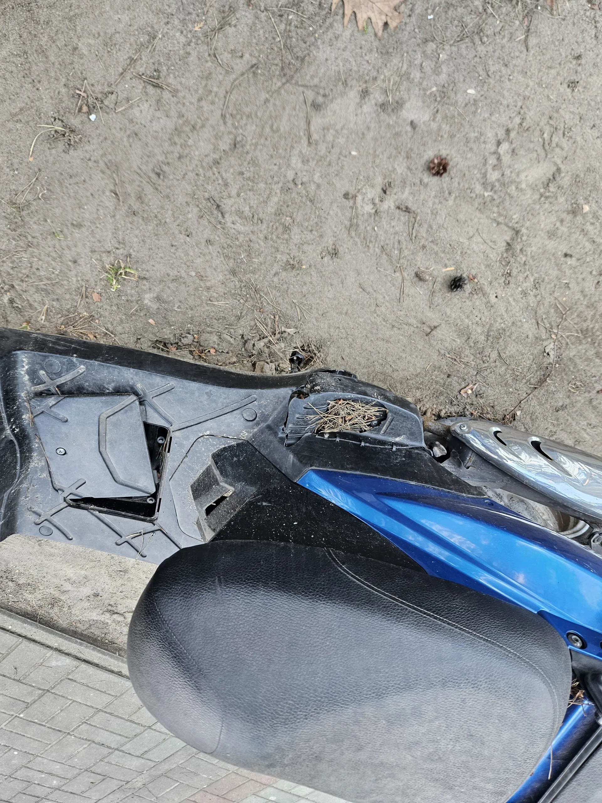 Zbliżenie na niebiesko-czarny skuter. Widoczne czarne siedzenie, uszkodzona czarna plastikowa część z suchą trawą lub słomą wewnątrz. Tło stanowi brudna ziemia z drobnymi roślinami i fragment chodnika z cegieł po lewej stronie. Część koła skutera widoczna u dołu.