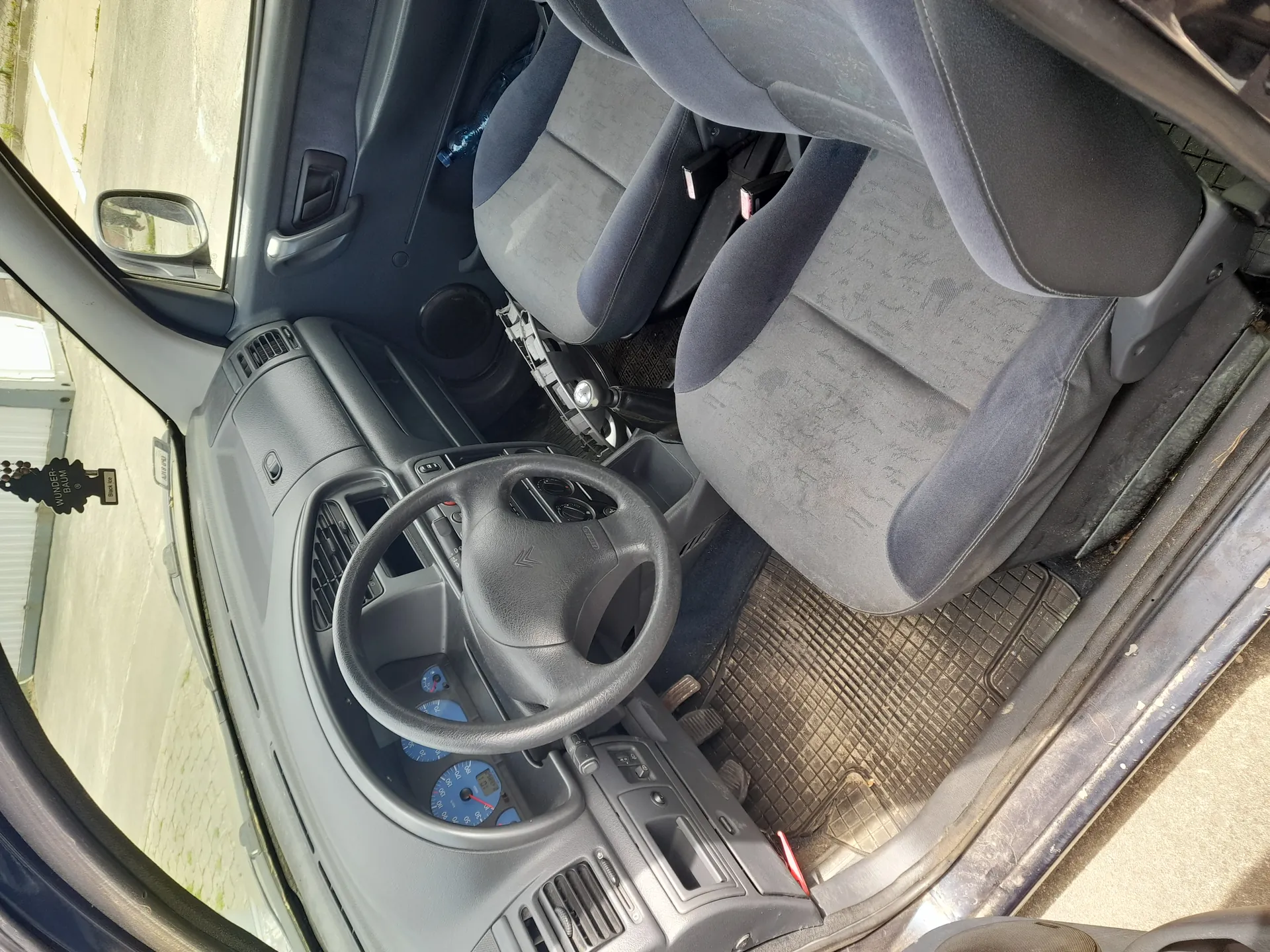 Wnętrze samochodu z widocznym kierownicą marki Citroën, szarymi tkaninowymi fotelami, deska rozdzielczą z niebieskimi wskaźnikami, otwartą drzwiami kierowcy i widokiem na ulicę oraz budynek poza pojazdem. Na podłodze czarne maty, a na fotelach widoczne ślady zużycia.