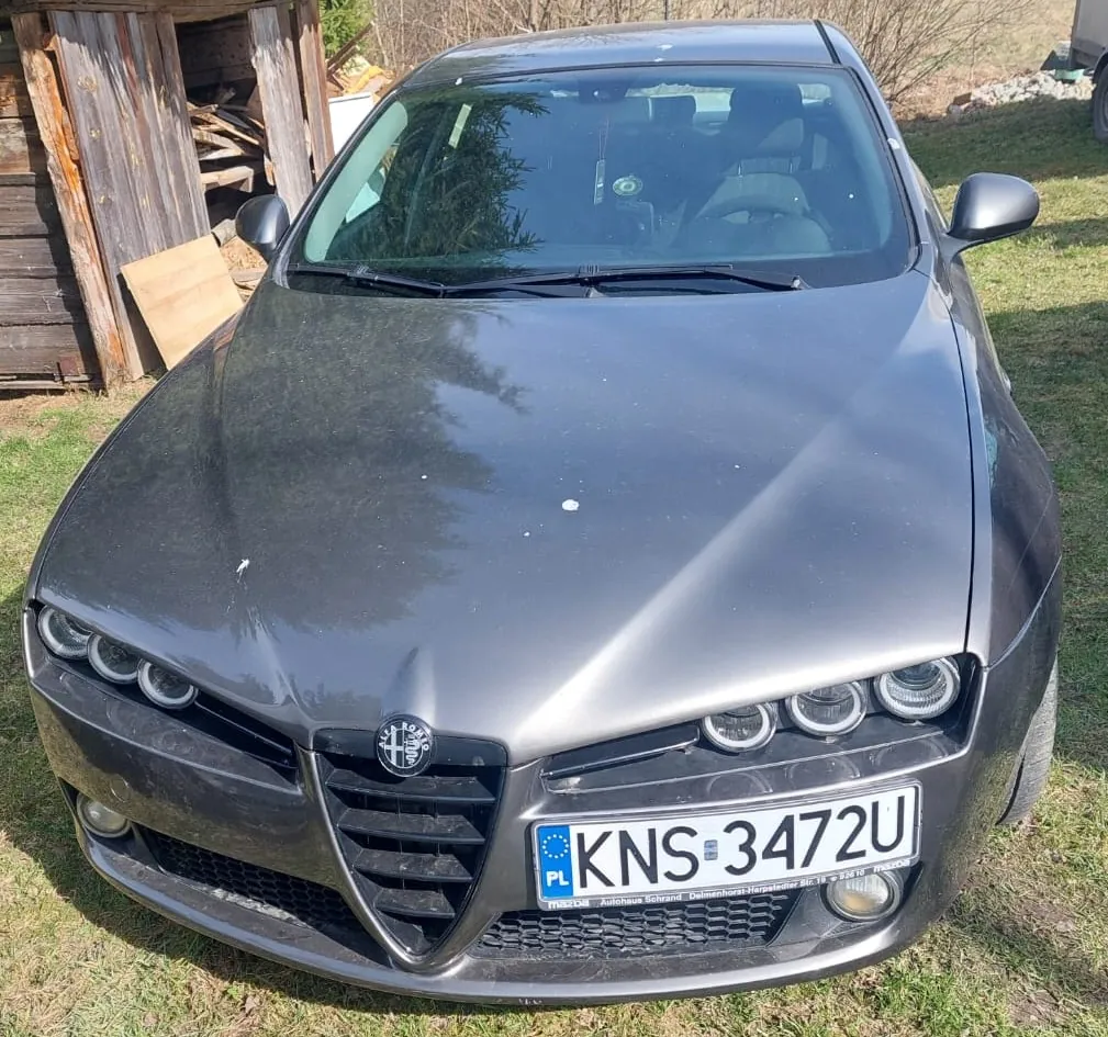 Szary samochód marki Alfa Romeo widziany od przodu, zaparkowany na trawie obok drewnianej szopy. Pojazd ma polską tablicę rejestracyjną o numerze KNS 3472U. Na masce widoczne są drobne zanieczyszczenia i odpryski lakieru.