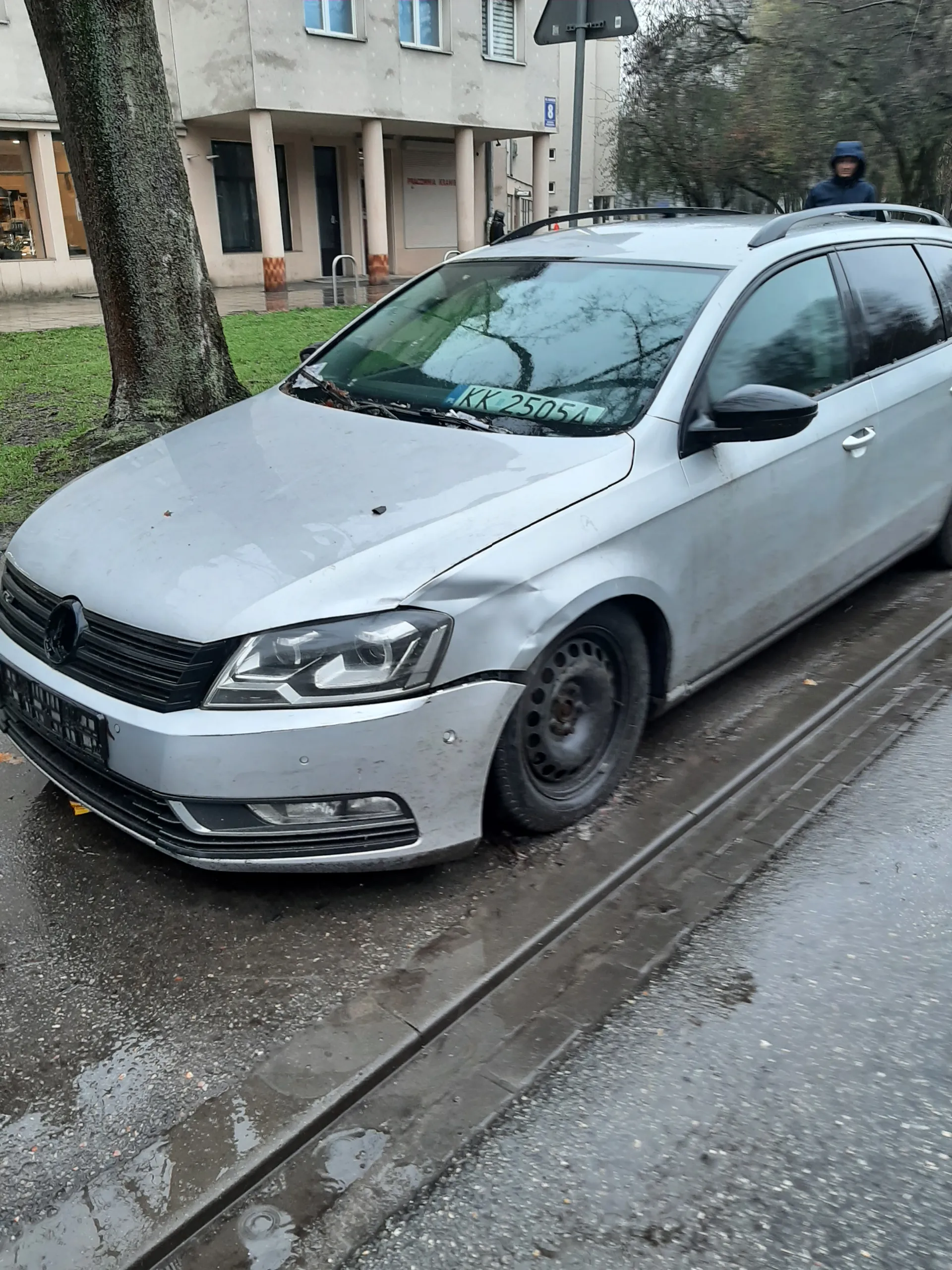 Srebrny samochód z widocznym uszkodzeniem przedniej prawej części. Tablica rejestracyjna KK 2505A. W tle budynek z kolumnami, drzewo i osoba w niebieskiej bluzie z kapturem. Mokra droga i chodnik.