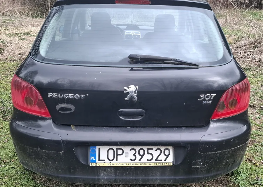 Tył czarnego samochodu Peugeot 307 HDI zaparkowanego na trawie. Widoczne są czerwone światła tylna, logo Peugeota, napisy 'PEUGEOT' po lewej stronie i '307 HDI' po prawej stronie. Tablica rejestracyjna PL LOP 39529 z żółtym pasem zawierającym tekst 'CZEŚCI DO AUT FRANCUSKICH tel. 22 783 37 88'. W tle sucha trawa i krzewy. Na szybie tylnej naklejka z napisem 'AT'.