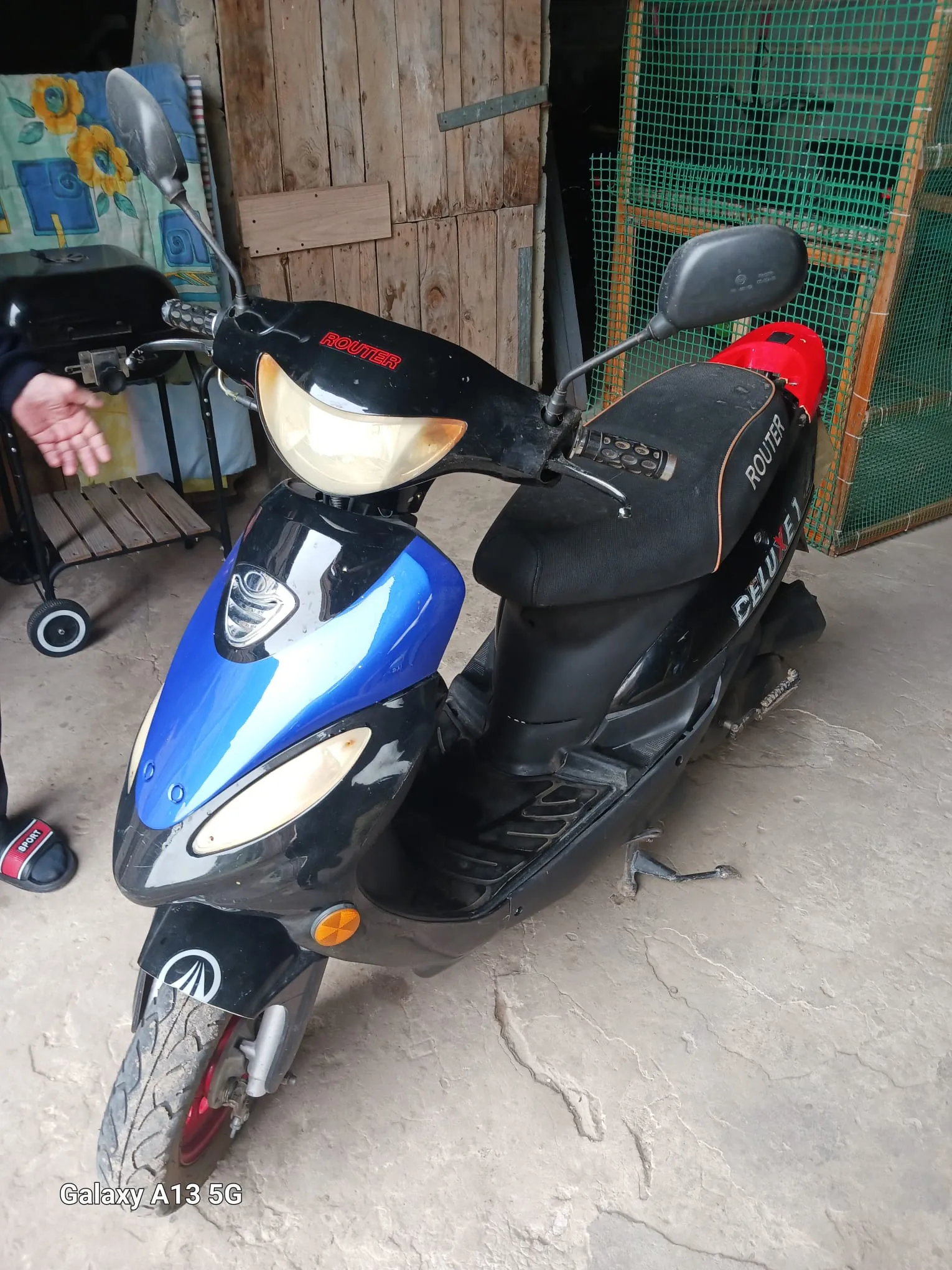 Czarno-niebieski skuter marki ROUTER z napisem DELUXE na boku, stojący na betonowej powierzchni. Na przedniej części skutera widoczny czerwony napis ROUTER, a na siedzeniu biały napis ROUTER. W tle drewniana brama, zielona siatka ogrodzeniowa oraz wózek z czarnym grillu. Po lewej stronie widoczna dłoń osoby i fragment buta z czerwonym napisem. W lewym dolnym rogu obrazu znajduje się napis Galaxy A13 5G.