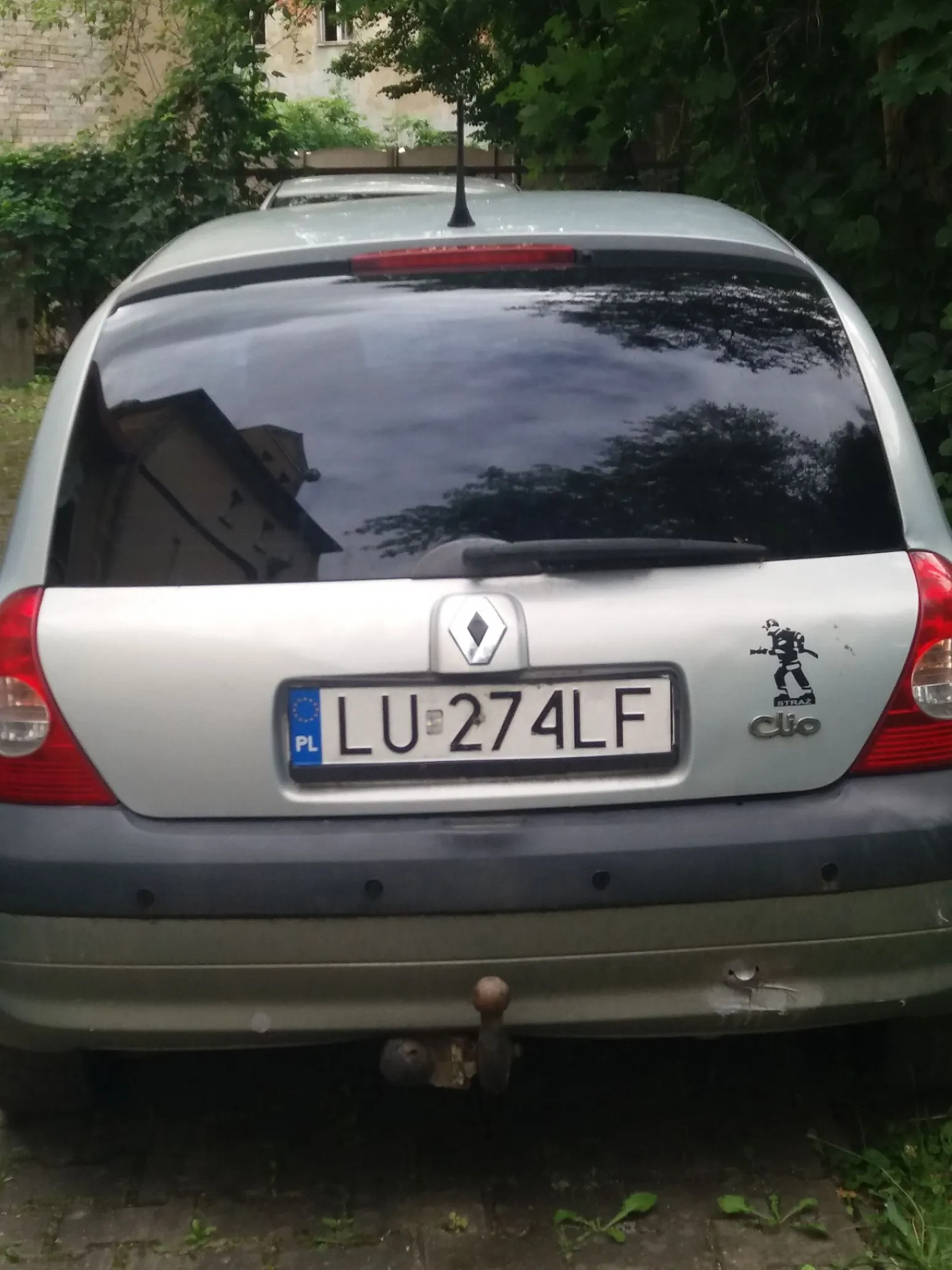 Tylny widok srebrnego samochodu marki Renault Clio. Na klapie bagażnika znajduje się polska tablica rejestracyjna o numerze LU 274LF oraz logo Renault. Po prawej stronie tablicy widnieje napis 'Clio' oraz naklejka z sylwetką postaci. Samochód posiada hak holowniczy. W tle widoczna jest zieleń i fragment budynku.