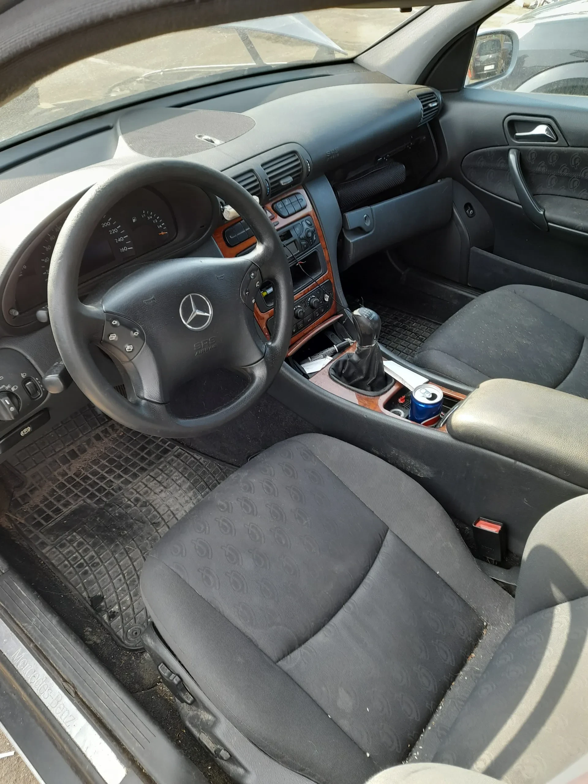 Wnętrze samochodu Mercedes-Benz z widocznym kierownicą z logo marki, czarnymi siedzeniami z powtarzającym się wzorem, centralną konsolą z przyciskami i dźwignią zmiany biegów. W uchwycie na napoje znajduje się niebieska puszka. Deska rozdzielcza z drewnianą obudową, brudne dywaniki podłogowe. Otwarte drzwi kierowcy z widocznym lusterkiem bocznym i napisem "Mercedes-Benz" na progu drzwi. W tle fragment zewnętrznej części pojazdu.