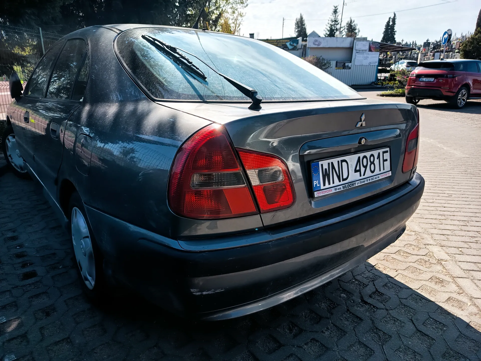 Z tyłu widoczny ciemnoszary samochód marki Mitsubishi z tablicą rejestracyjną PL WND 4981F. Poniżej tablicy napis: TOYOTA W-wa, WŁOCHY ul. Stawy 16 22 863 44 44 TOYOTA. Samochód zaparkowany na brukowanej powierzchni. W tle czerwony SUV, budynki, drzewa i niebieski znak drogowy. Słoneczny dzień, widoczne cienie na ziemi.
