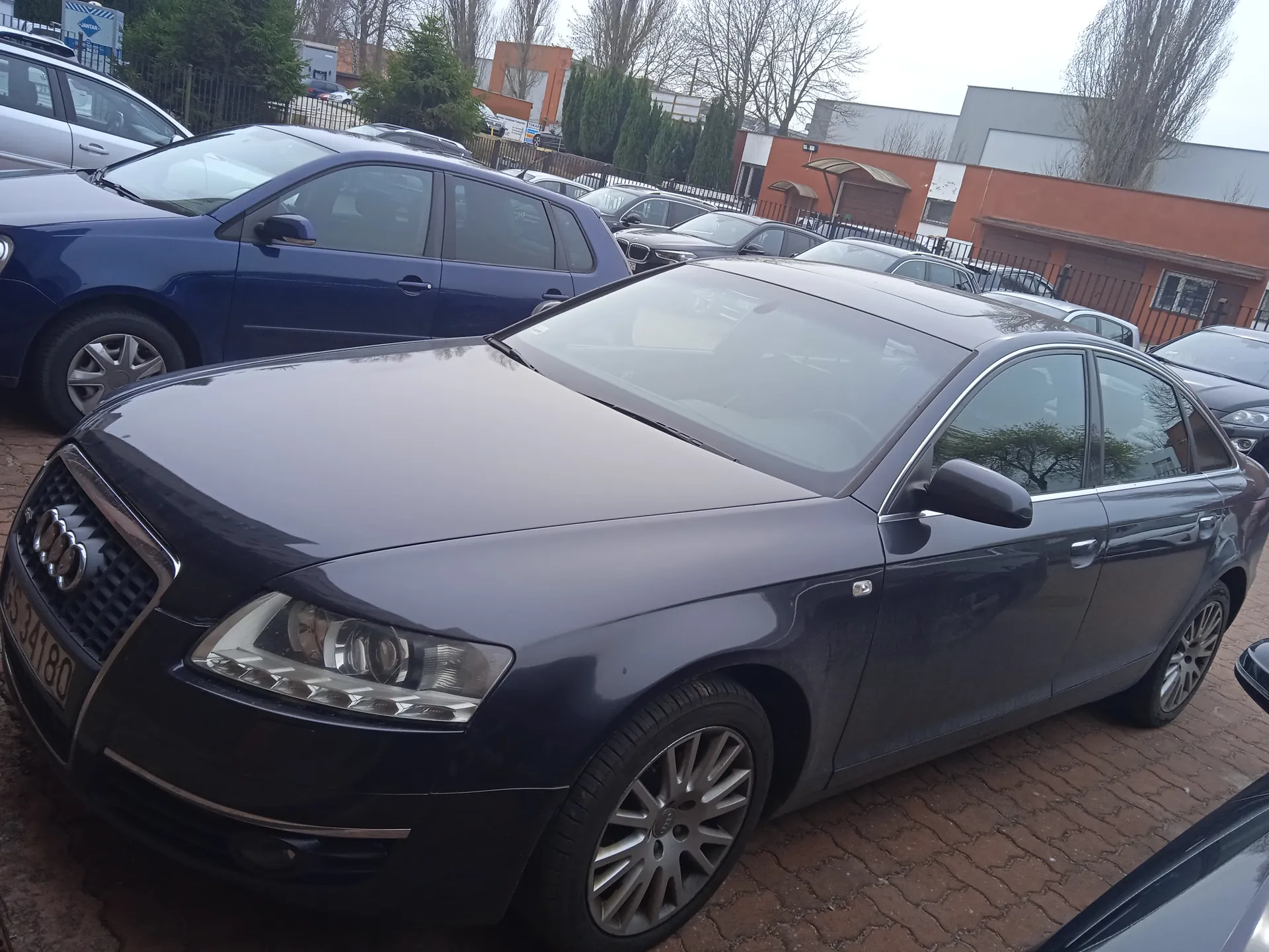 Ciemnoszary samochód Audi A6 z tablicą rejestracyjną BS 34180 zaparkowany na ceglanym placu parkingowym. W tle widoczne są inne pojazdy, w tym niebieski samochód osobowy, budynki przemysłowe z pomarańczowymi i szarymi elewacjami, drzewa bez liści oraz metalowe ogrodzenie. Niebo jest pochmurne.