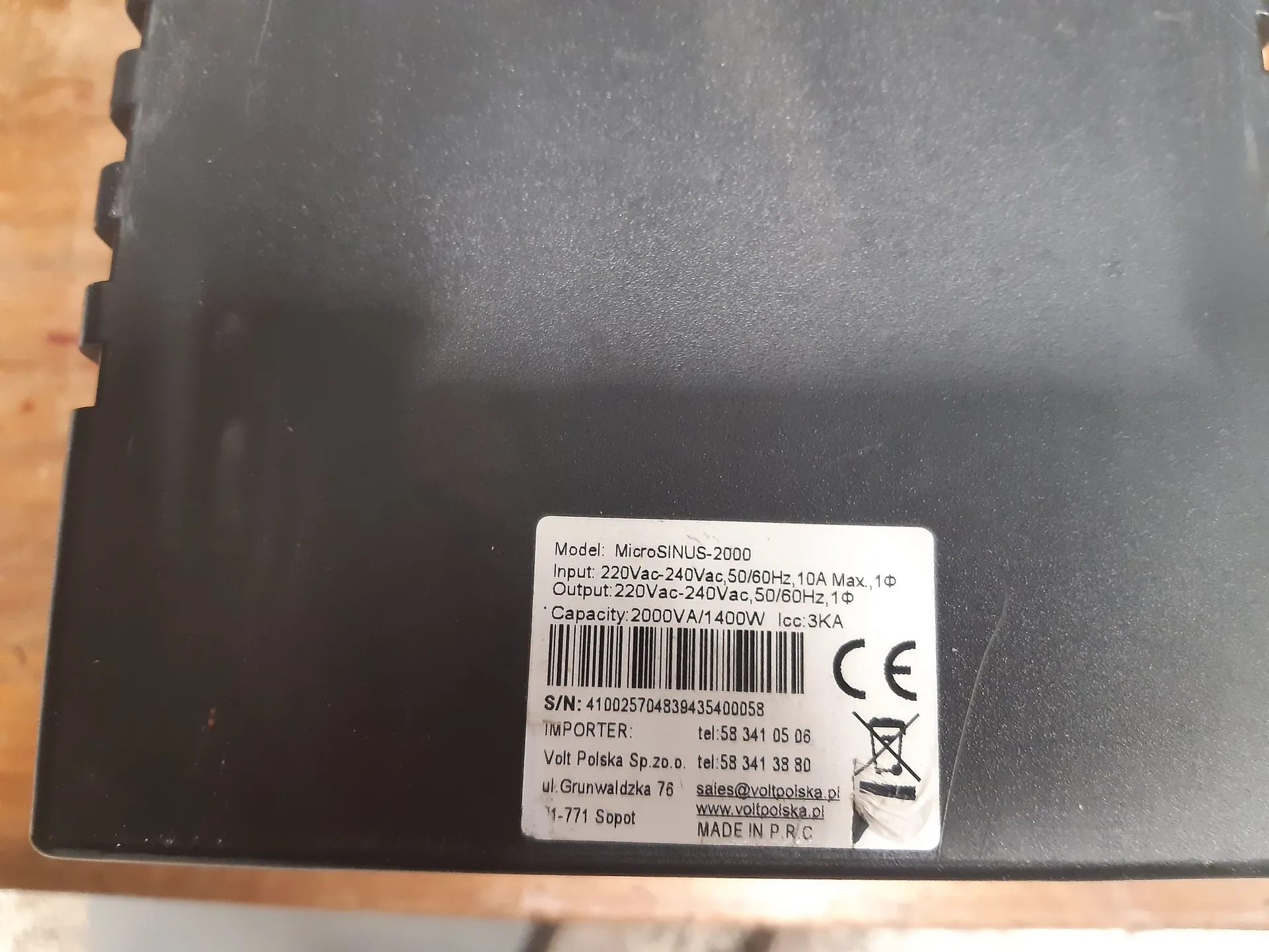 Zbliżenie na czarną obudowę urządzenia elektronicznego z naklejoną białą etykietą techniczną. Na etykiecie znajduje się tekst: 'Model: MicroSINUS-2000', 'Input: 220Vac-240Vac, 50/60Hz, 10A Max., 1Φ', 'Output: 220Vac-240Vac, 50/60Hz, 1Φ', 'Capacity: 2000VA/1400W Icc 3KA'. Widoczny jest kod kreskowy, numer seryjny 'S/N: 410025704839435400058' oraz dane importera: 'Volt Polska Sp. z o.o. ul. Grunwaldzka 76 81-771 Sopot', numery telefonów, adres e-mail 'sales@voltpolska.pl' i strona internetowa 'www.voltpolska.pl'. Na dole widnieje napis 'MADE IN P.R.C' oraz oznaczenia CE i symbol przekreślonego kosza na śmieci.
