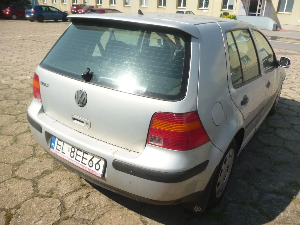 Srebrny samochód marki Volkswagen Golf zaparkowany na kostce brukowej. Widok z tyłu i z boku pojazdu. Samochód ma polską tablicę rejestracyjną o numerze EL 8EE66. W tle widoczne są inne zaparkowane samochody oraz fragment budynku.