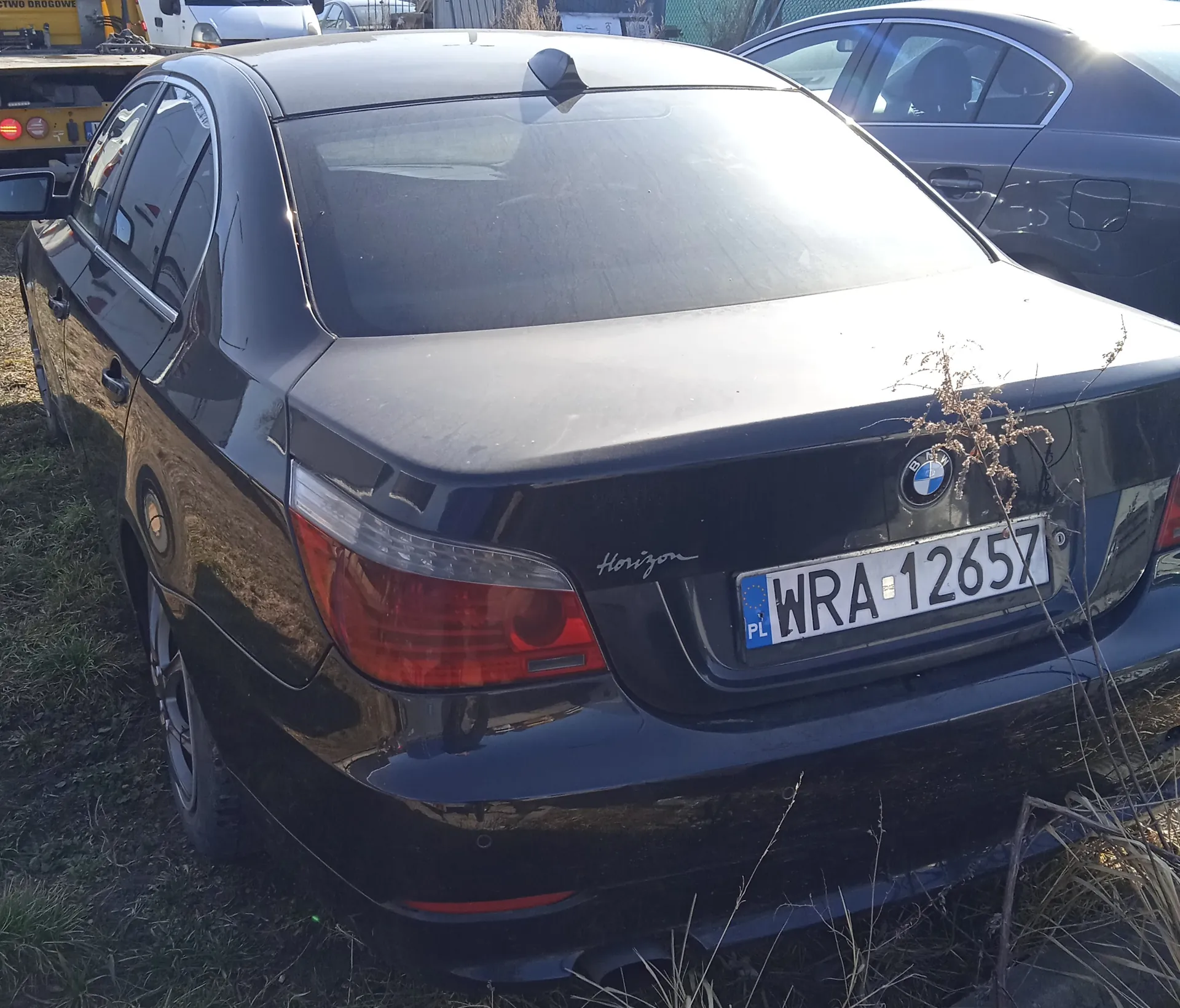 Czarny samochód marki BMW z widokiem od tyłu. Na bagażniku widoczny napis 'Horizon' oraz logo BMW. Tablica rejestracyjna z polskim kodem 'PL' i numerem 'WRA-12657'. Samochód zaparkowany na trawie, w tle inne pojazdy, w tym żółty holownik i ciemny samochód osobowy. Słoneczne oświetlenie powoduje połysk na tylnej szybie.