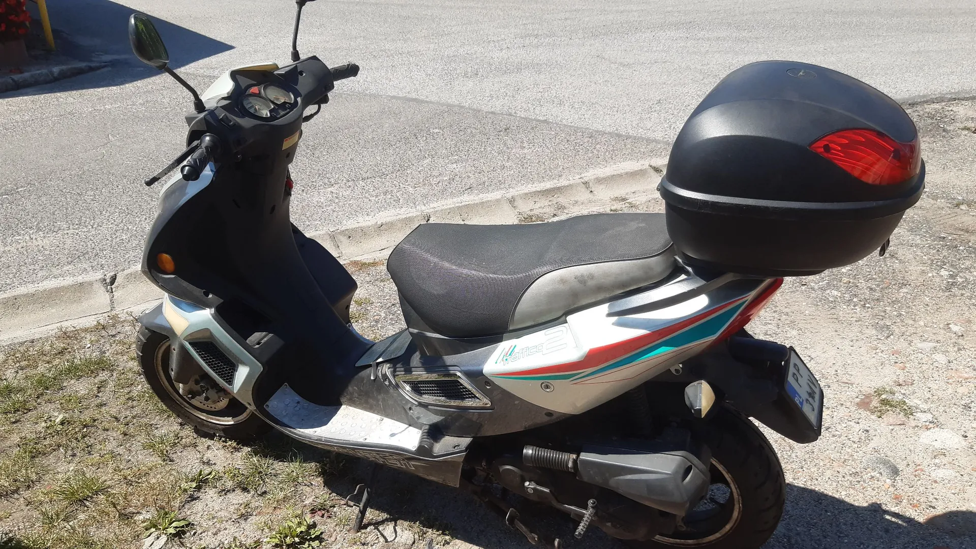Srebrno-szary skuter zaparkowany na poboczu drogi. Pojazd posiada czarne siedzenie oraz czarny kufer zamontowany z tyłu. Na boku skutera widoczne są kolorowe pasy w kolorach czerwonym i niebieskim oraz napis 'Techno'. Skuter jest ustawiony bokiem do obiektywu.
