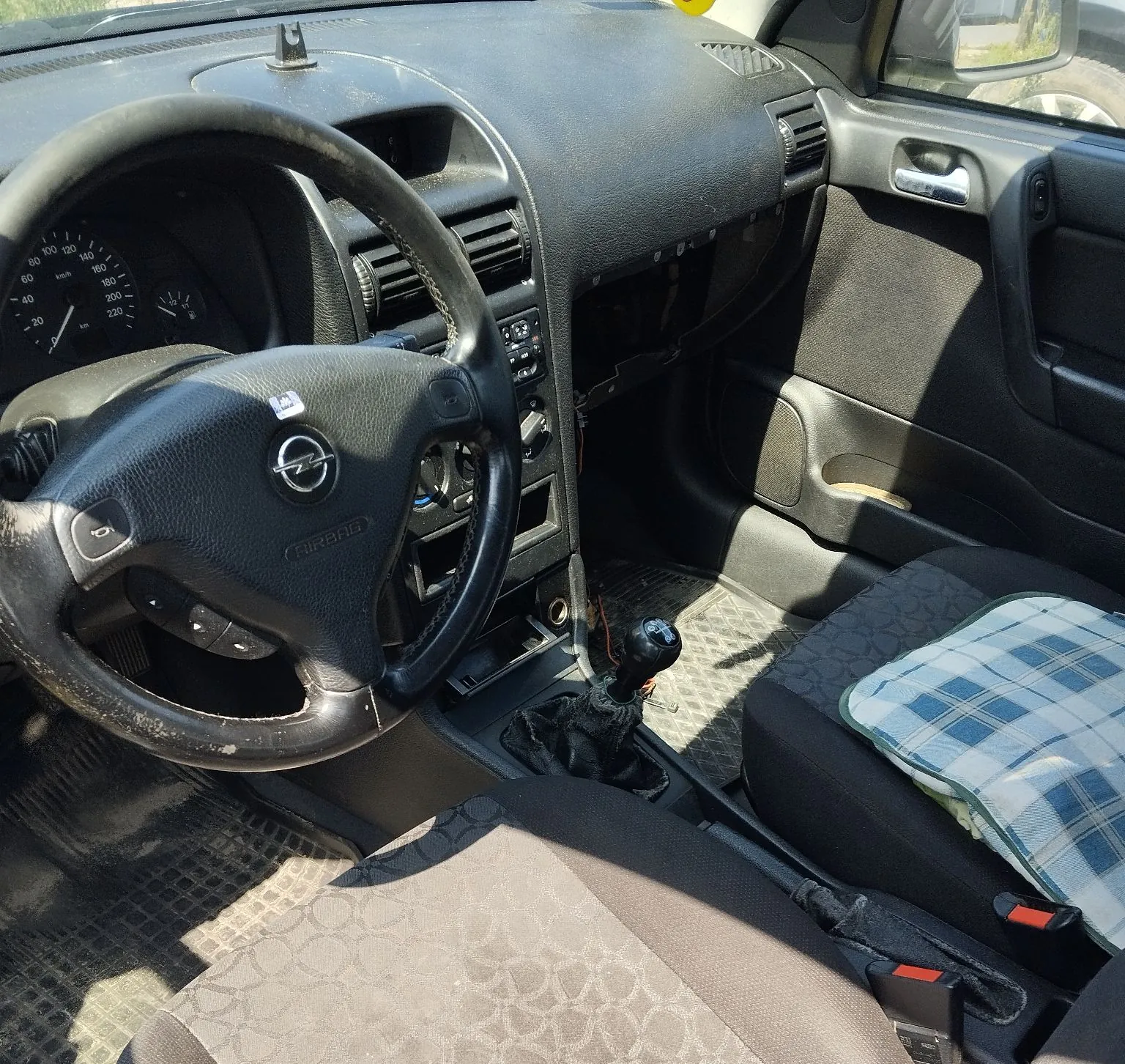 Wnętrze samochodu marki Opel, widok z perspektywy kierowcy. Widoczna czarna kierownica z logo producenta i napisem AIRBAG, deska rozdzielcza z licznikiem prędkości, lewarek zmiany biegów oraz dwa przednie fotele. Pasażerski fotel jest częściowo przykryty niebiesko-białą tkaniną w kratkę. W konsoli środkowej brakuje niektórych elementów wykończenia.