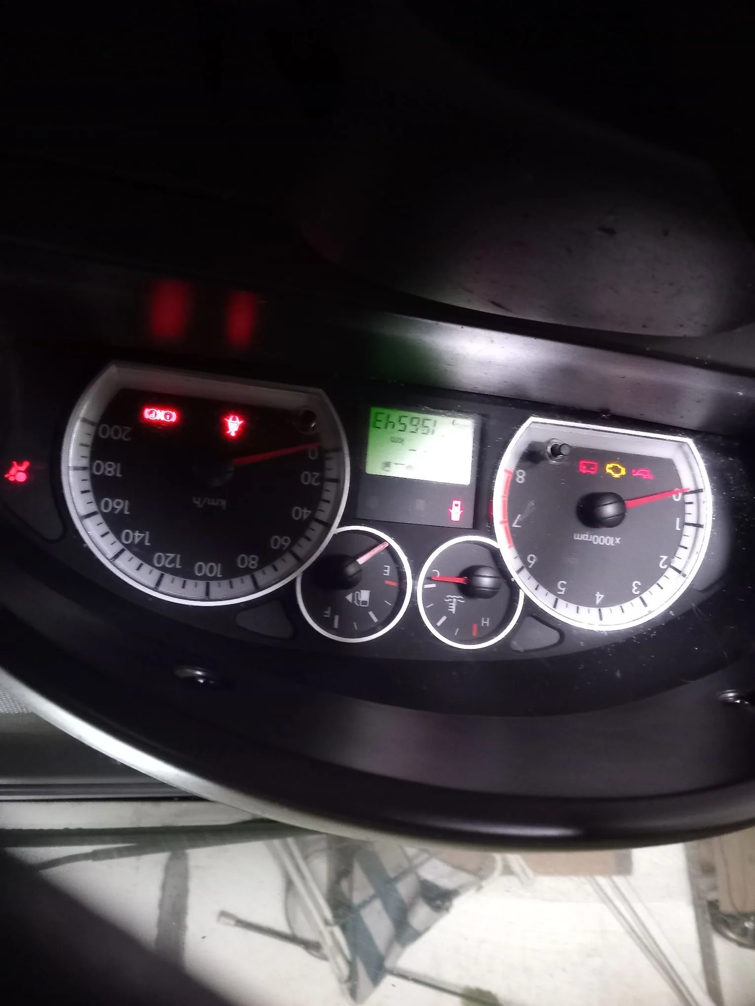 Zdjęcie deski rozdzielczej samochodu z licznikiem prędkości, obrotomierzem i wskaźnikami paliwa oraz temperatury. Licznik prędkości wskazuje 0 km/h, a obrotomierze również 0. Cyfrowy licznik przebiegu pokazuje wartość 156543. Na panelu świecą się czerwone kontrolki ostrzegawcze.
