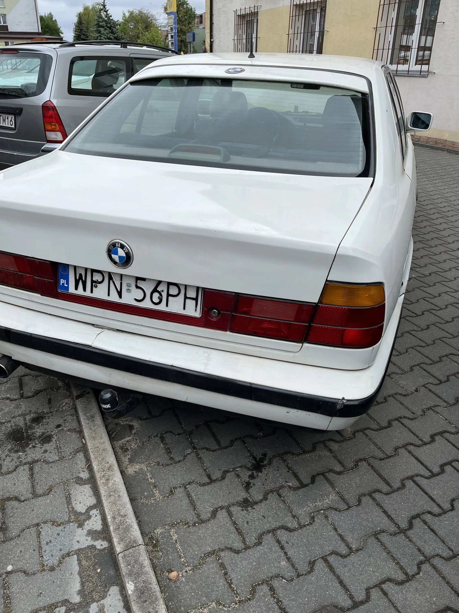 Biały samochód marki BMW z widocznym tyłem, w tym czerwonymi światłami tylnymi i tablicą rejestracyjną WPN 56PH z niebieskim pasem PL. Samochód zaparkowany na brukowanej nawierzchni, w tle częściowo widoczny inny pojazd oraz budynki z oknami i drzewami. Tablica rejestracyjna umieszczona pionowo, co wskazuje na nieprawidłową orientację obrazu.