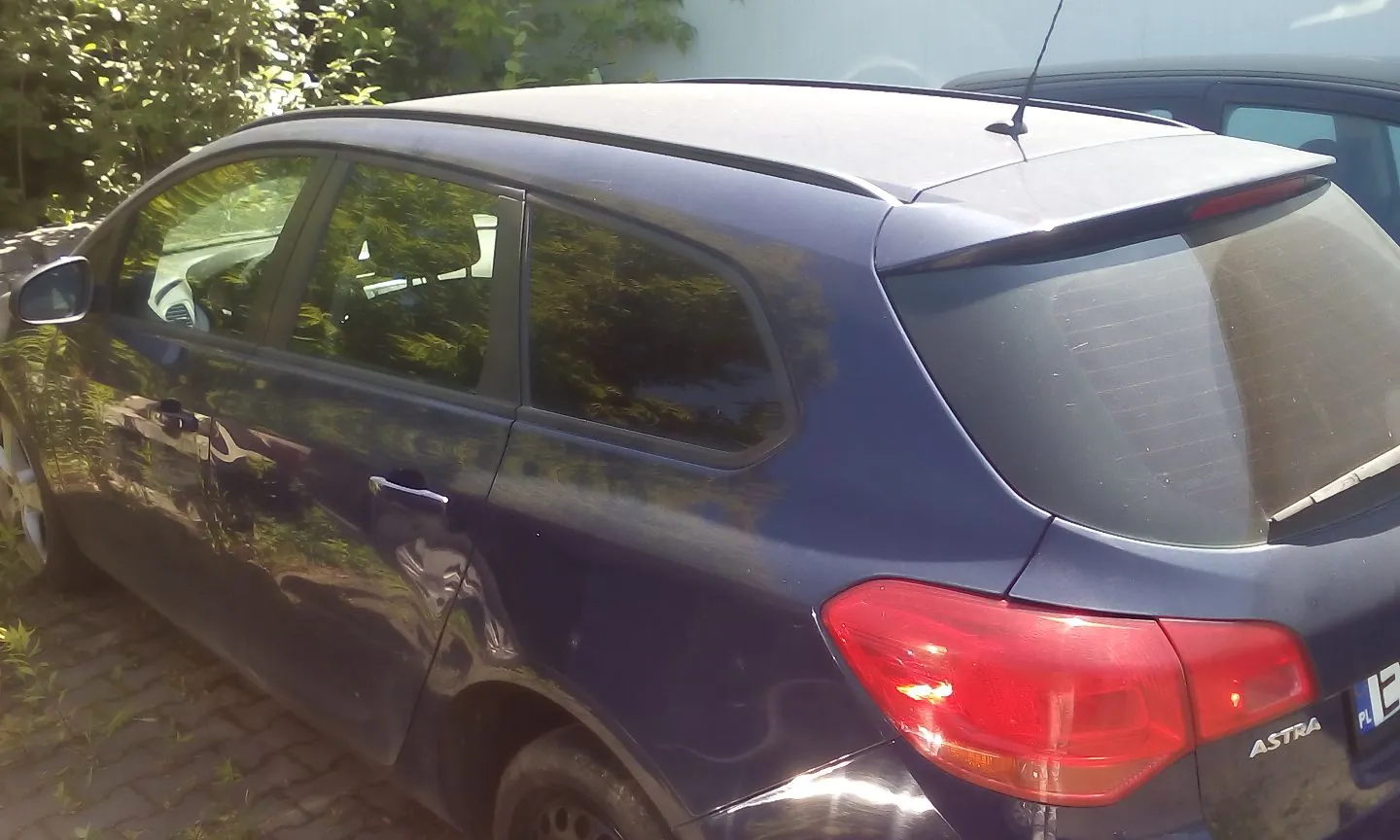 Widok z boku i z tyłu ciemnoniebieskiego samochodu marki Opel Astra w nadwoziu kombi. Pojazd jest zaparkowany na kostce brukowej obok zielonych krzewów. Na tylnej klapie widoczny jest napis 'ASTRA' oraz fragment tablicy rejestracyjnej z oznaczeniem 'PL'.