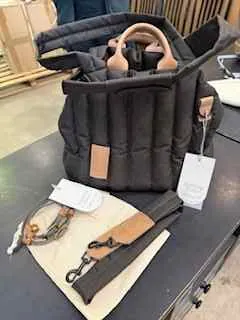 Czarna pikowana kurtka z brązowym skórzany elementem na klatce piersiowej i białą etykietą. Obok kurtki znajduje się beżowa torba z brązowym paskiem i metalowym zamkiem oraz brązowy skórzany pasek z metalowymi elementami. Tło stanowi ciemna powierzchnia stołu i częściowo widoczne pudełka z kartonu.