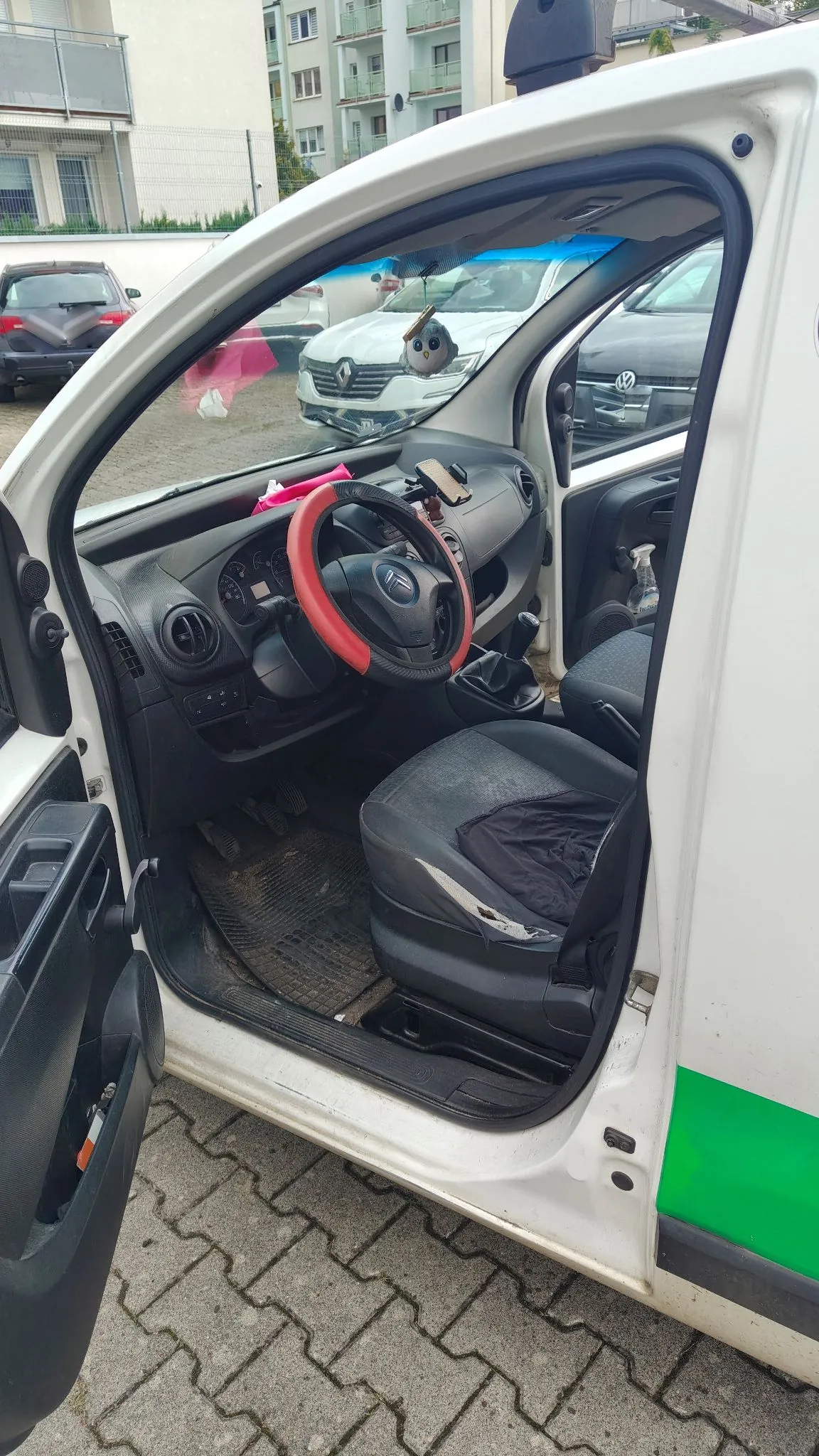 Widok z otwartych drzwi kierowcy do wnętrza białego samochodu dostawczego. Wewnątrz widoczna jest czarna kierownica z czerwonym pokrowcem, ciemnoszara tapicerka fotela kierowcy oraz deska rozdzielcza. Na lusterku wstecznym wisi mała ozdoba w kształcie białej postaci. Samochód zaparkowany jest na kostce brukowej, w tle widoczne są inne zaparkowane auta oraz wielopiętrowy budynek mieszkalny.