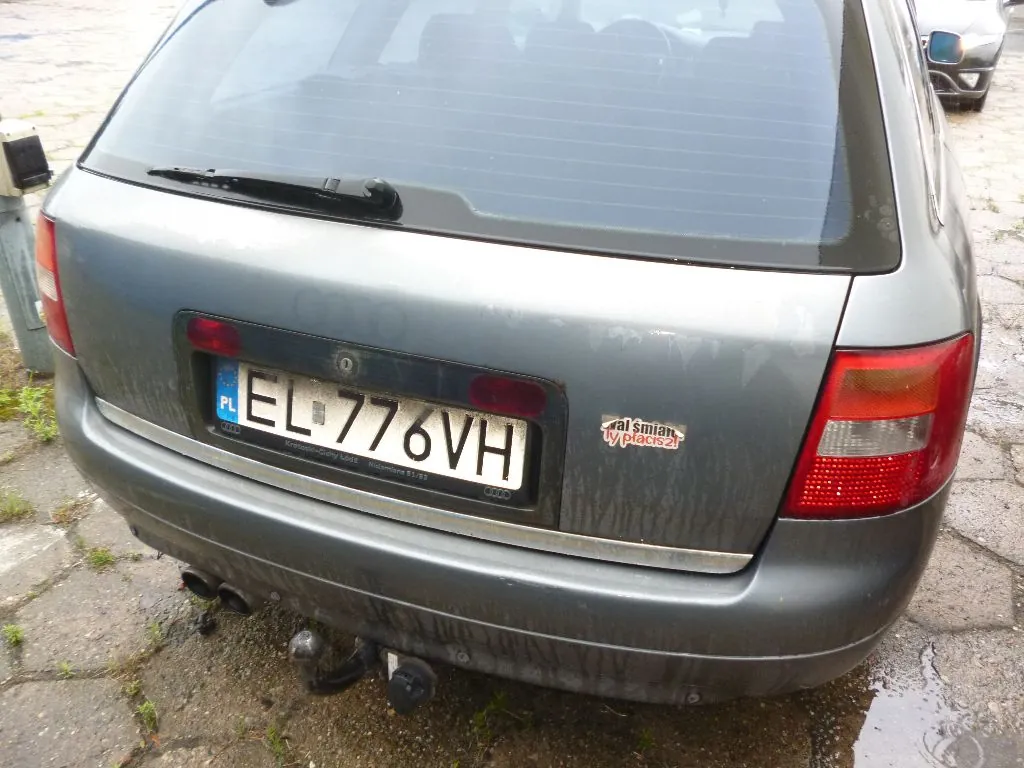 Tylny widok szarego samochodu osobowego. Widoczna jest klapa bagażnika z tablicą rejestracyjną o numerze EL 776VH, z niebieskim paskiem z napisem PL po lewej stronie. Po prawej stronie tablicy znajduje się mała naklejka z tekstem „al Śmiań ty placez2!”. Samochód ma podwójny wydech po lewej stronie oraz zamontowany hak holowniczy. Podwozie i dolna część zderzaka są zabrudzone błotem.