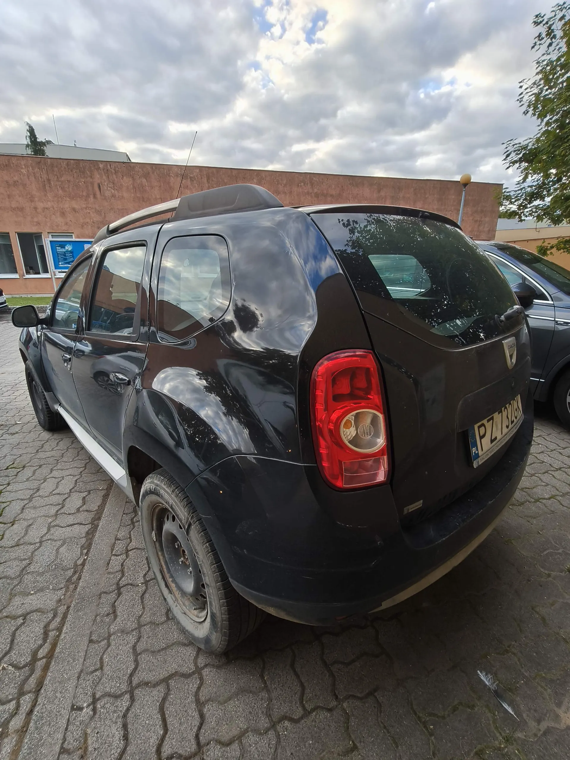 Czarny samochód marki Dacia z tyłu i z boku, zaparkowany na kostce brukowej. Pojazd posiada relingi dachowe i polską tablicę rejestracyjną o numerze PZ 732GK. W tle widoczny jest brązowy budynek oraz zachmurzone niebo.