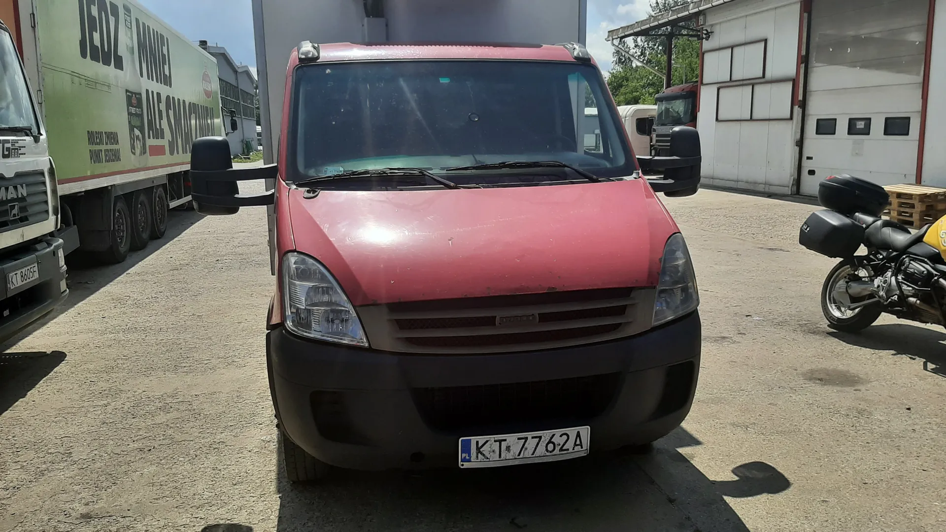 Czerwony samochód dostawczy Iveco z tablicą rejestracyjną KT 7762A (PL) na pierwszym planie. Po lewej stronie zielony naczepa z napisem JEDZ MNIEJ ALE SMACZNIEJ oraz mniejszym tekstem ROZKŁAD JAZDY PUNKT JEDZENIA. Po prawej stronie motocykl z czarnymi kuframi. Tło: budynek z białymi garażami, inne pojazdy i drzewa. Asfaltowy plac parkingowy.