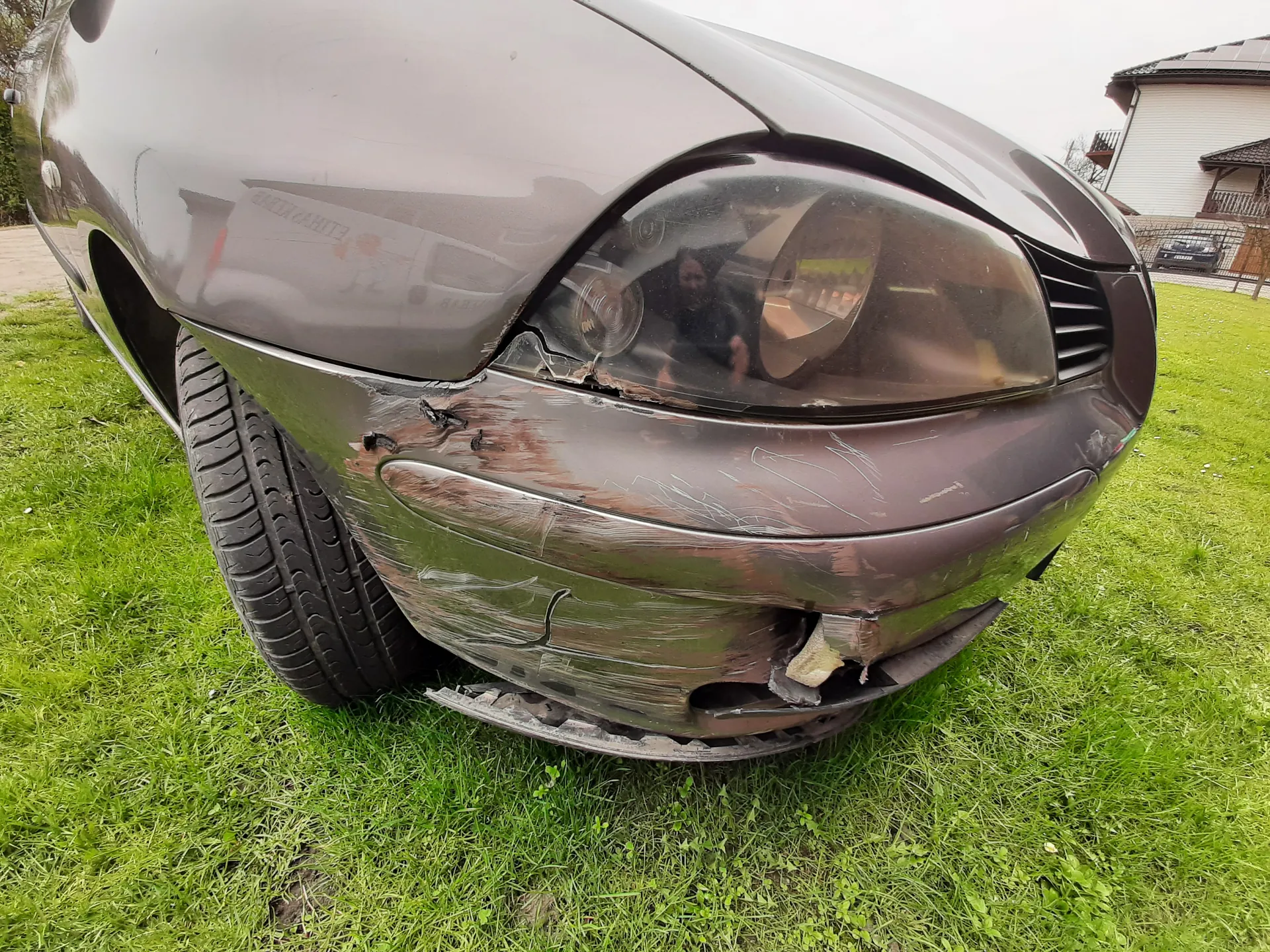 Przednia część uszkodzonego ciemnoszarego samochodu osobowego z pękniętym zderzakiem i uszkodzonym reflektorem. Samochód stoi na trawie, w tle widoczny biały dom z ciemnym dachem.
