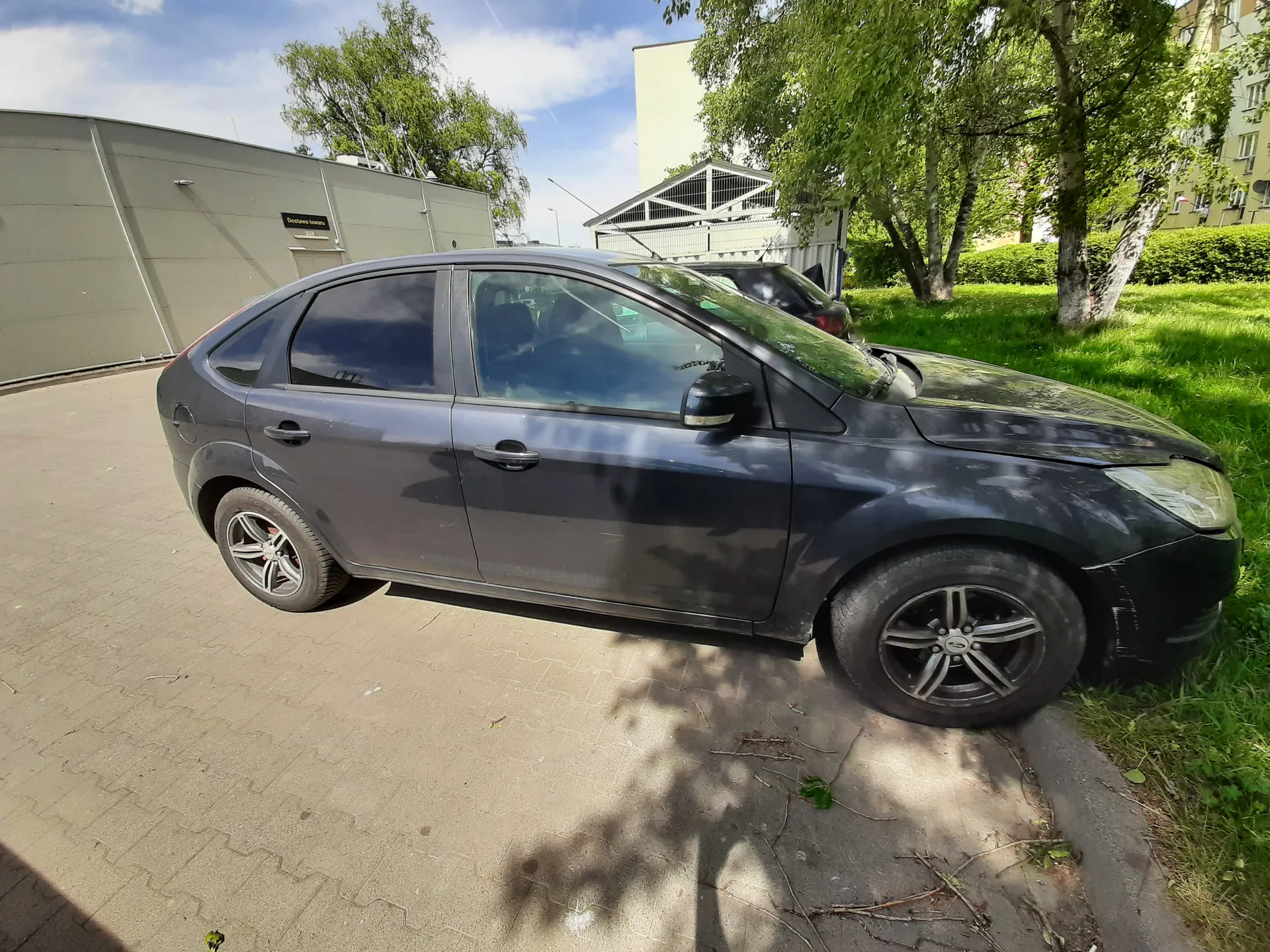 Ciemnoszary samochód osobowy typu hatchback zaparkowany na brukowanej powierzchni. Po lewej stronie widoczny budynek z tablicą o napisie "Dostawa towaru", za samochodem drzewa, trawnik oraz fragmenty innych budynków. Na tle niebo z chmurami. W tle częściowo widoczny inny samochód.