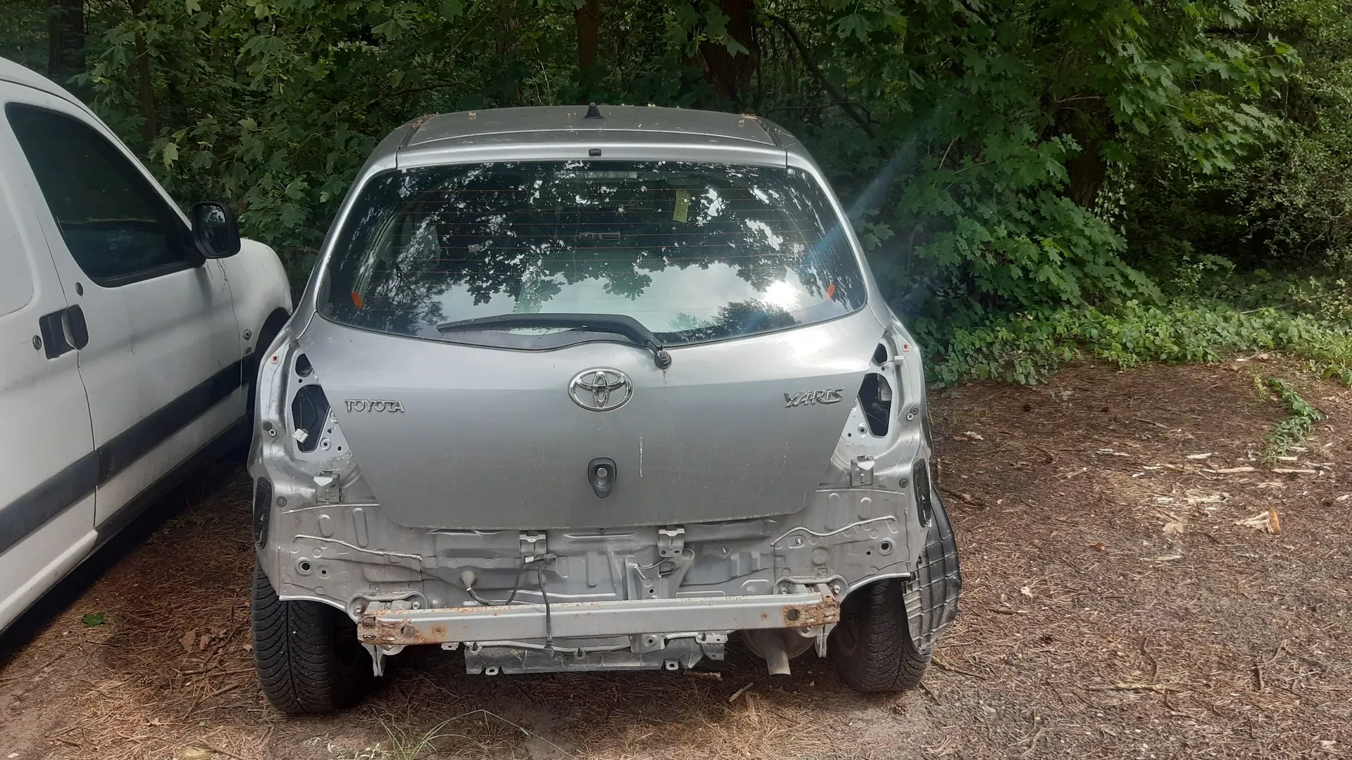Srebrny samochód marki Toyota Yaris z uszkodzonym tyłem, zaparkowany na leśnej drodze obok białego vana. Samochód nie posiada tylnego zderzaka, odsłaniając metalową belkę wzmacniającą i elementy konstrukcyjne nadwozia. Na klapie bagażnika widoczne są napisy 'TOYOTA' oraz 'YARIS' oraz logo marki.
