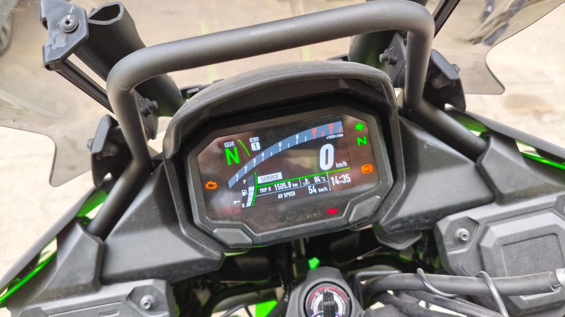 Zbliżenie na cyfrowy wyświetlacz motocykla. Na ekranie widoczna jest prędkość 0 km/h, bieg neutralny (N), obrotomierz, temperatura 84 °C, godzina 14:35 oraz przebieg TRIP B 1505.9 km. Wyświetlacz jest obramowany czarną, plastikową obudową, a nad nim znajduje się czarny metalowy stelaż ochronny.