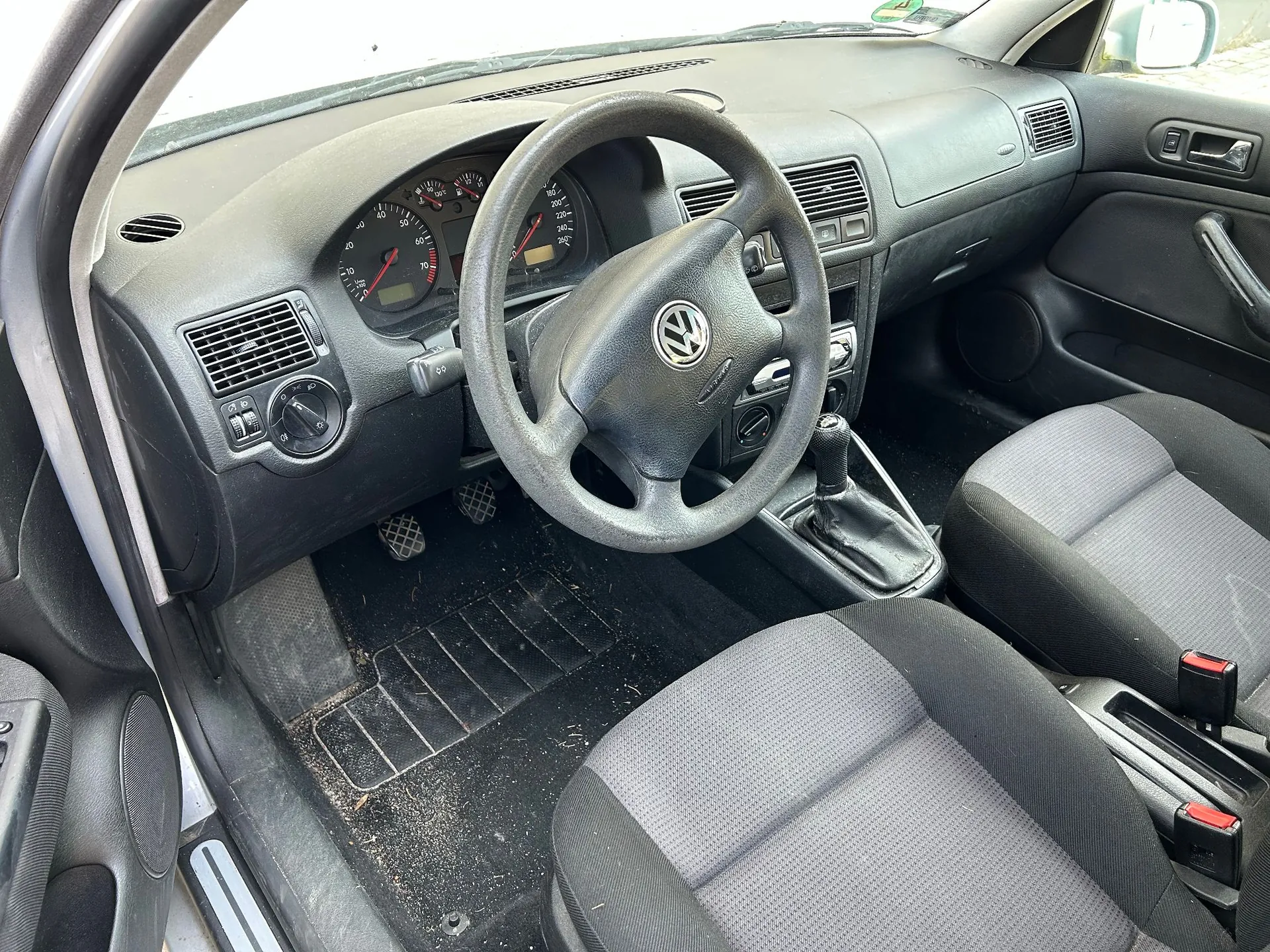 Wnętrze samochodu z widokiem na deskę rozdzielczą, kierownicę z logo Volkswagena, zegary analogowe oraz dwa przednie fotele z szarym materiałem. Widoczna jest konsola środkowa, pedały oraz czarna wykładzina na podłodze.