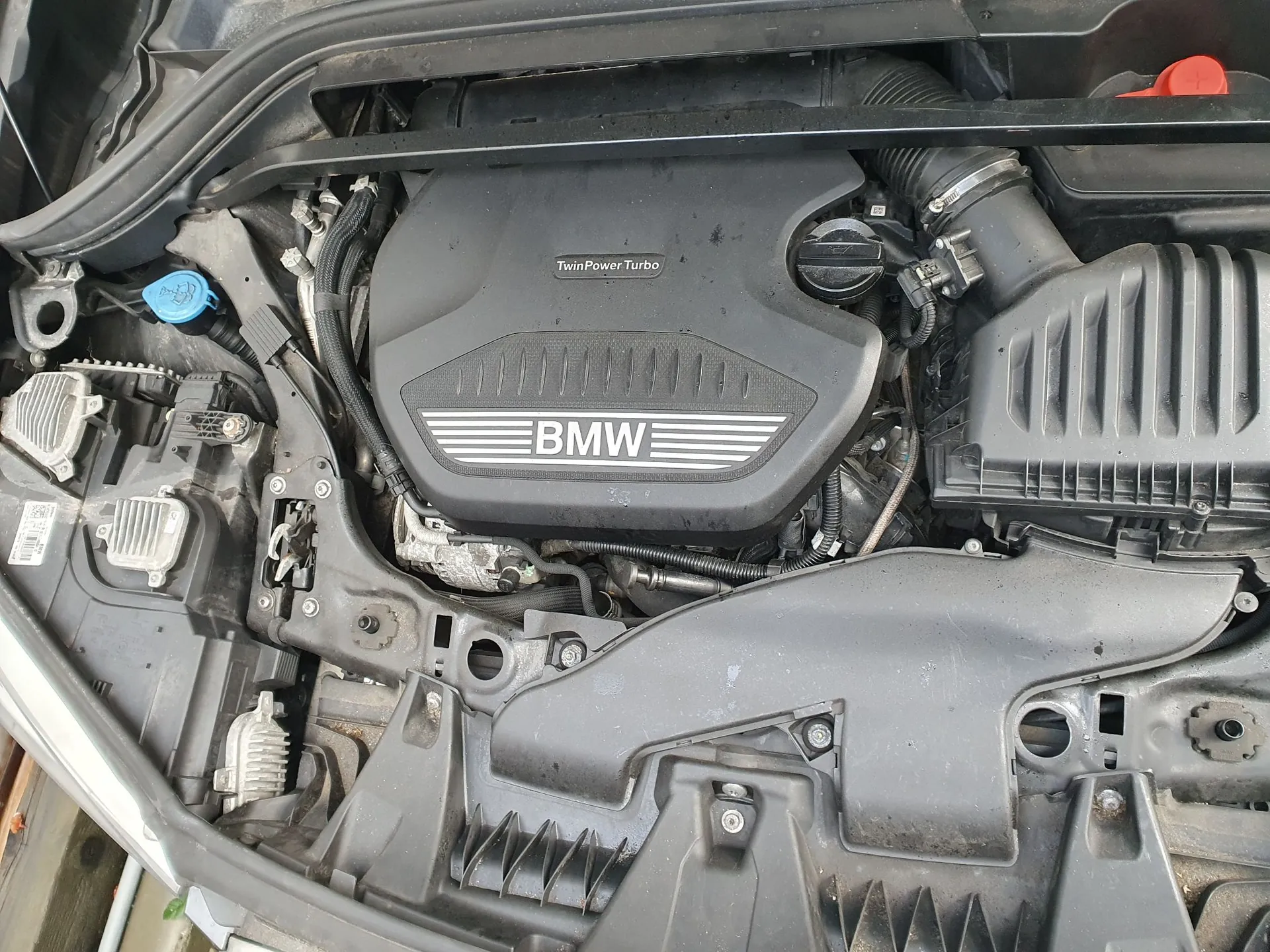 Widok z góry na komorę silnika samochodu marki BMW. Centralnie znajduje się czarna plastikowa osłona silnika z napisem 'TwinPower Turbo' oraz dużym logo 'BMW'. Po prawej stronie widoczny jest obudowa filtra powietrza, a po lewej niebieska zakrętka zbiornika płynu spryskiwaczy. W prawym górnym rogu znajduje się czerwona dźwignia lub zakrętka.