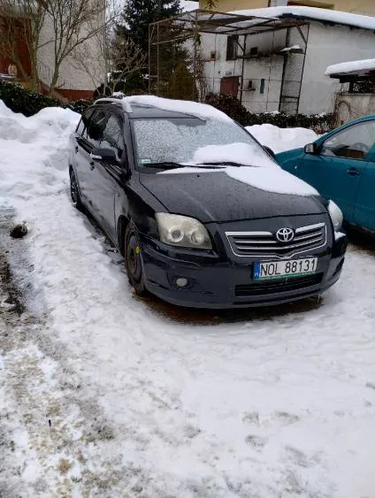 Samochód marki Toyota, kolor ciemnoszary, pokryty śniegiem, z widoczną tablicą rejestracyjną NOL 88131. Obok niego stoi niebieski samochód, również z śniegiem na dachu. Tło stanowią budynki mieszkalne, drzewo bez liści oraz konstrukcja z daszkiem. Całość otoczona śniegiem na ziemi z widocznymi śladami stóp.