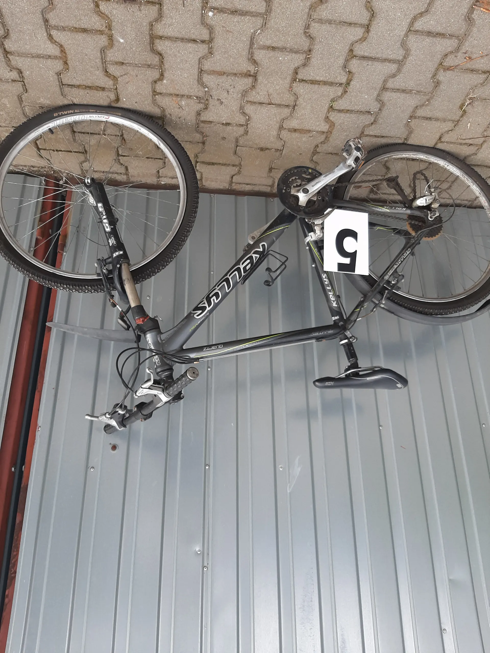 Czarny rower marki KELLYS oparty o szarą metalową bramę garażową z pionowymi listwami. Na ramie roweru widoczny biały napis KELLYS. Przy ramie zamocowana biała tabliczka z czarną cyfrą 5. Tło po prawej stronie to brukowana powierzchnia z kamiennymi płytami w kształcie litery U. Rower posiada czarne koła, srebrne elementy kierownicy oraz czarne siedzisko.