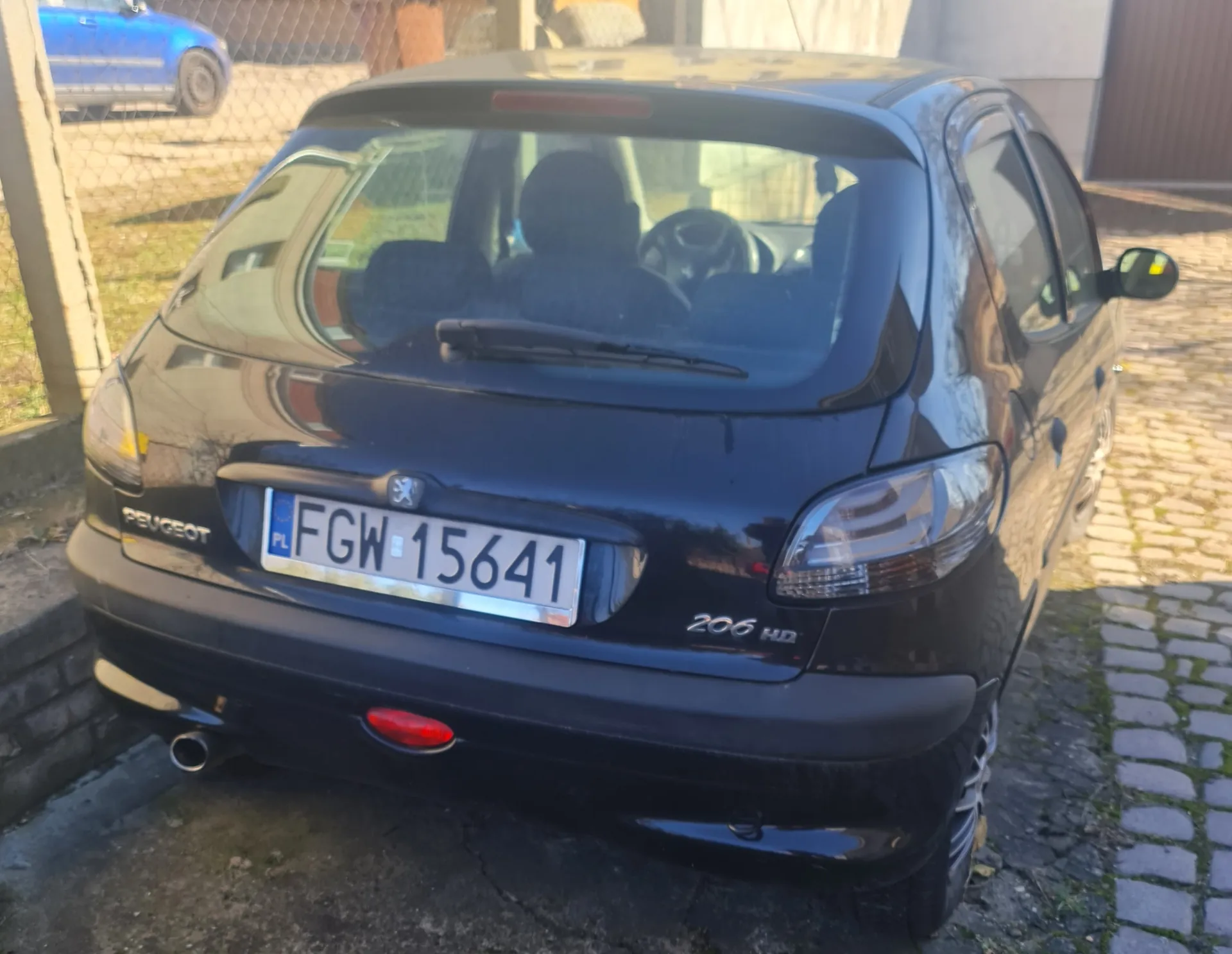 Czarny samochód Peugeot 206 HD widziany od tyłu, z tablicą rejestracyjną PL FGW 15641. Po lewej stronie widoczny jest napis 'PEUGEOT', a po prawej '206 HD'. Samochód zaparkowany na brukowanej powierzchni, w tle siatka ogrodzeniowa i fragment niebieskiego pojazdu. Na tylnej szybie widać sylwetki siedzeń i kierownicy.
