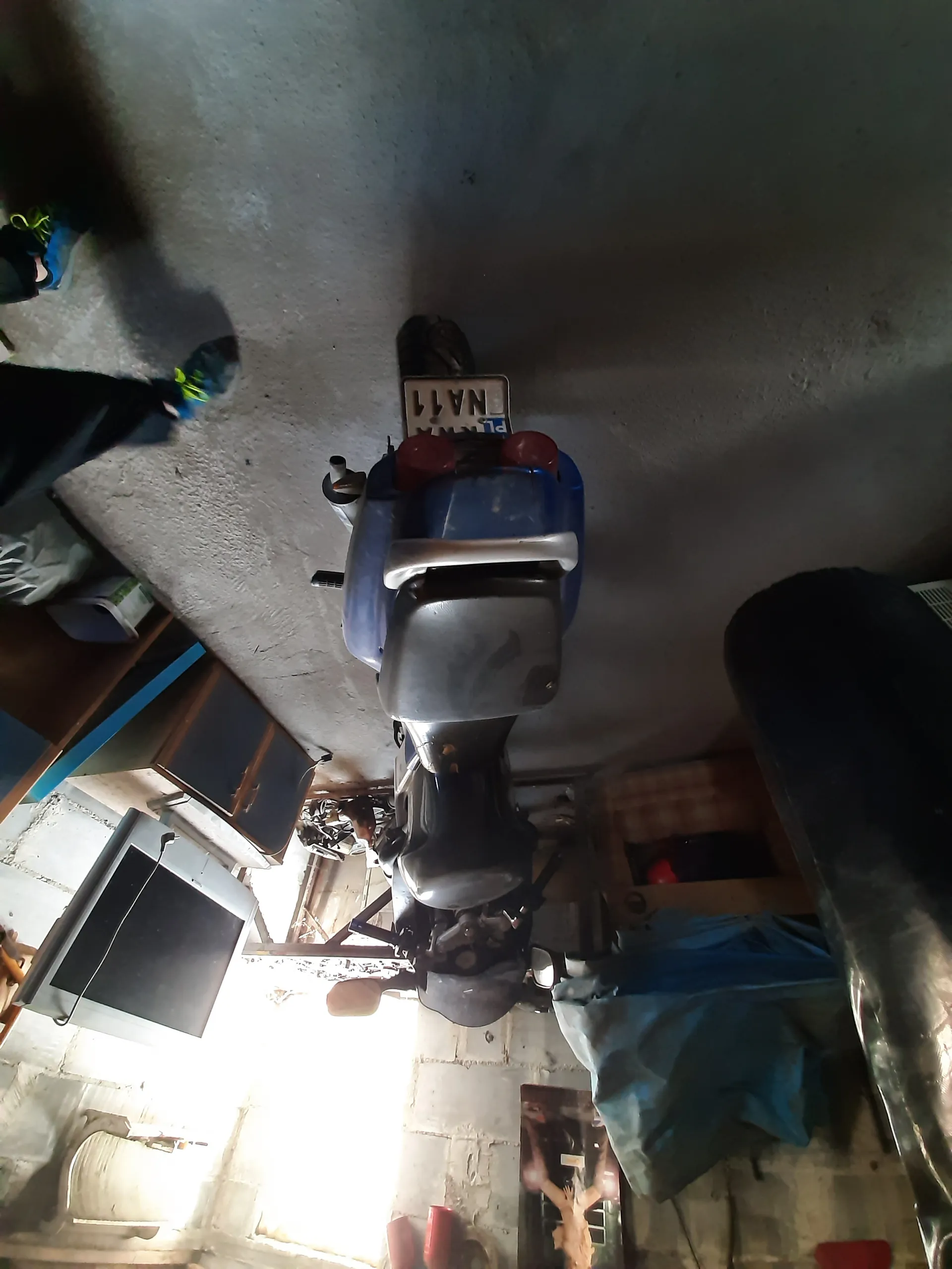 Niebieski skuter z tablicą rejestracyjną PL NA11 umieszczony w garażu lub warsztacie. W tle widoczne betonowe ściany, szafki, stary telewizor, osoba w ciemnych spodniach i niebieskich butach z zielonymi sznurkami oraz przedmioty przykryte niebieską folią. Po lewej stronie źródło światła wpadające przez otwór. Na podłodze betonowej leżą różne przedmioty, w tym czerwone pojemniki.