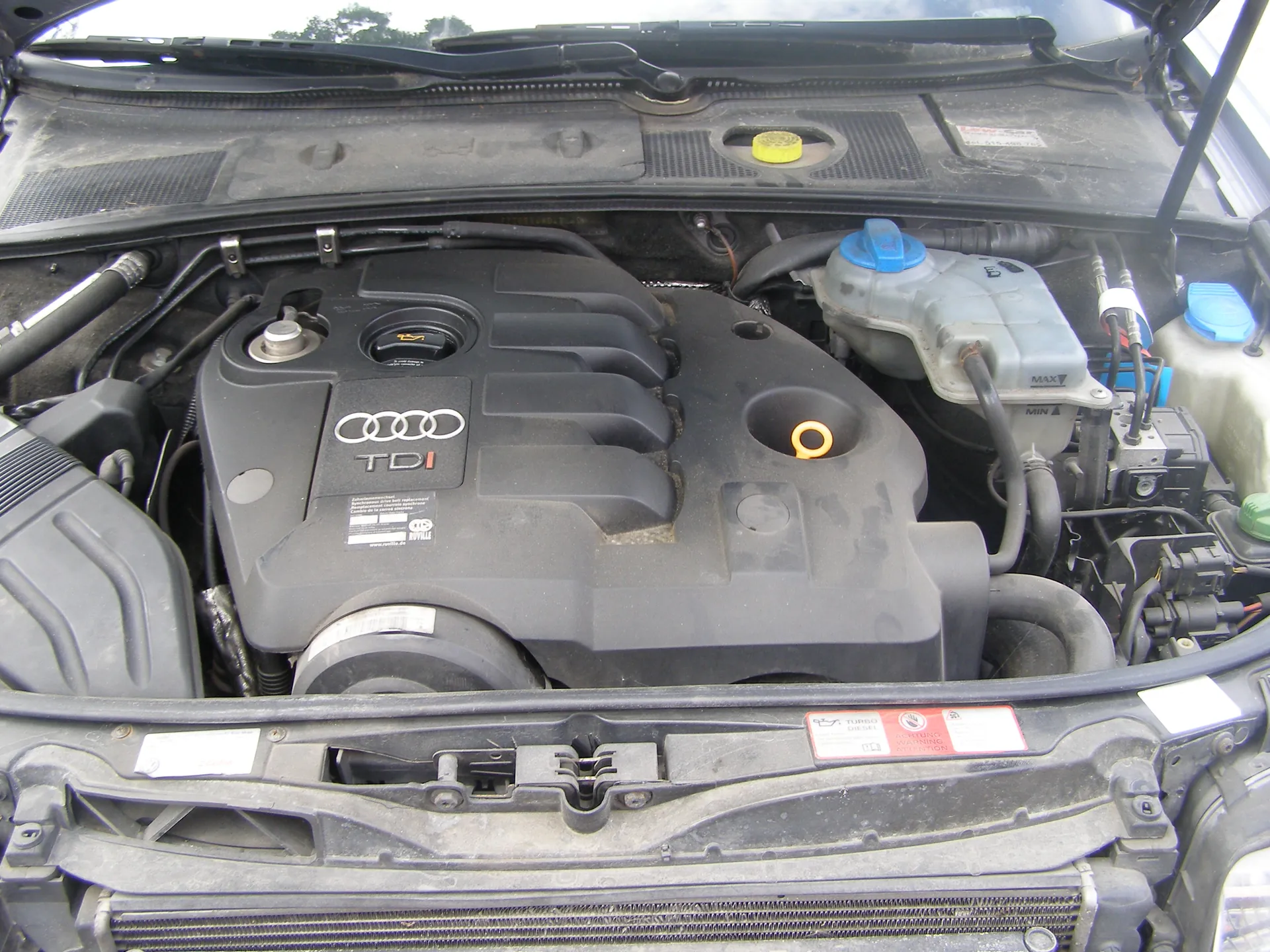 Otwarty kapota samochodu marki Audi z widocznym silnikiem TDI. Na pokrywie silnika umieszczono logo Audi (cztery połączone pierścienie) oraz napis "TDI". Wokół silnika znajdują się elementy takie jak żółta nakrętka, niebieskie nakrętki na zbiornikach płynów, przewody i etykiety. Na przedniej części komory silnikowej widoczna czerwono-biała etykieta z ostrzeżeniami.