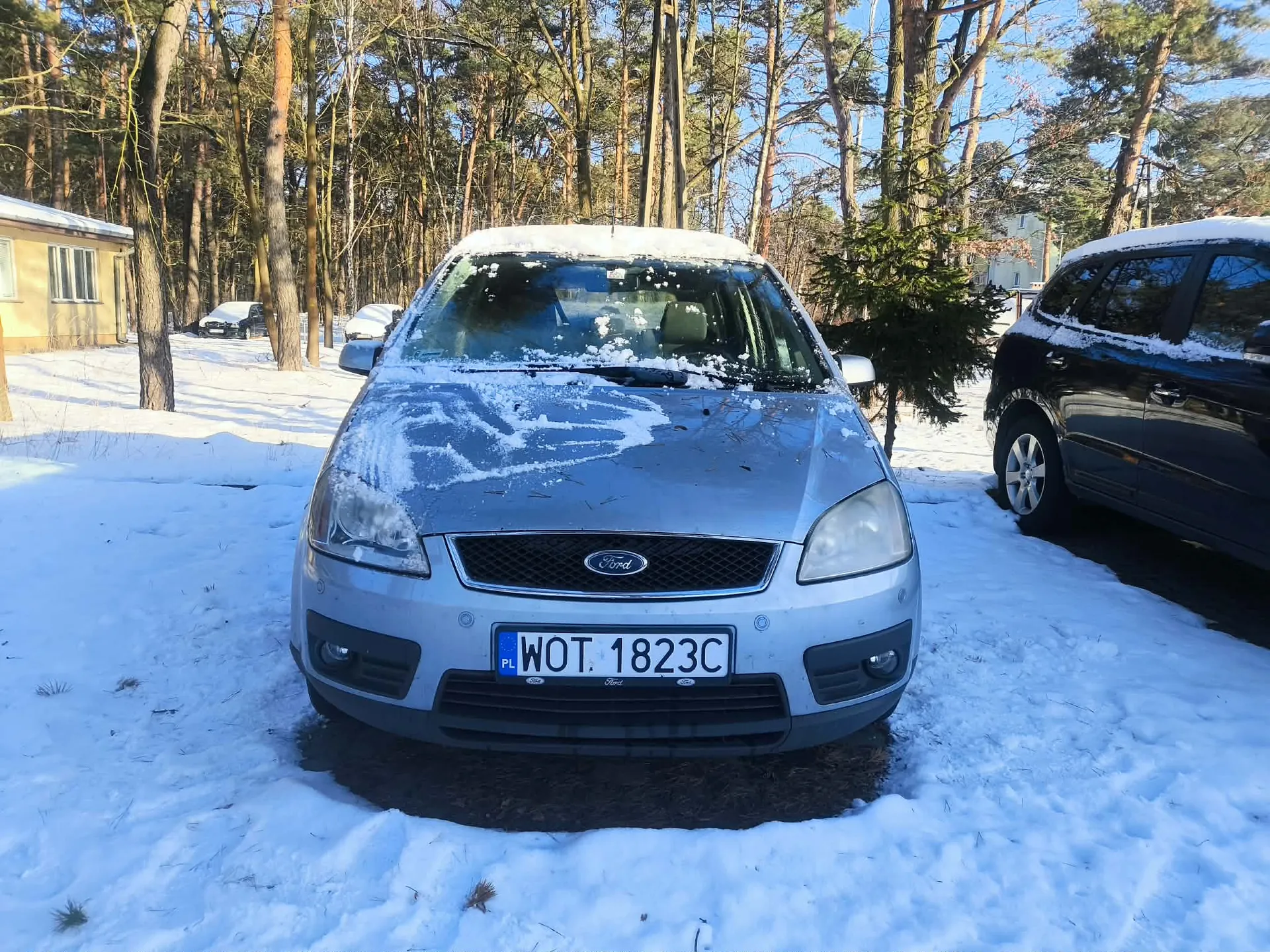 Srebrny samochód marki Ford z tablicą rejestracyjną WOT 1823C (PL) zaparkowany na śnieżnej powierzchni. Na masce i dachu pojazdu widoczny śnieg. Po prawej stronie czarny samochód, również pokryty śniegiem. Tło stanowią wysokie drzewa, domy i inne zaparkowane pojazdy. Całość otoczona śnieżnym krajobrazem w słoneczny dzień.